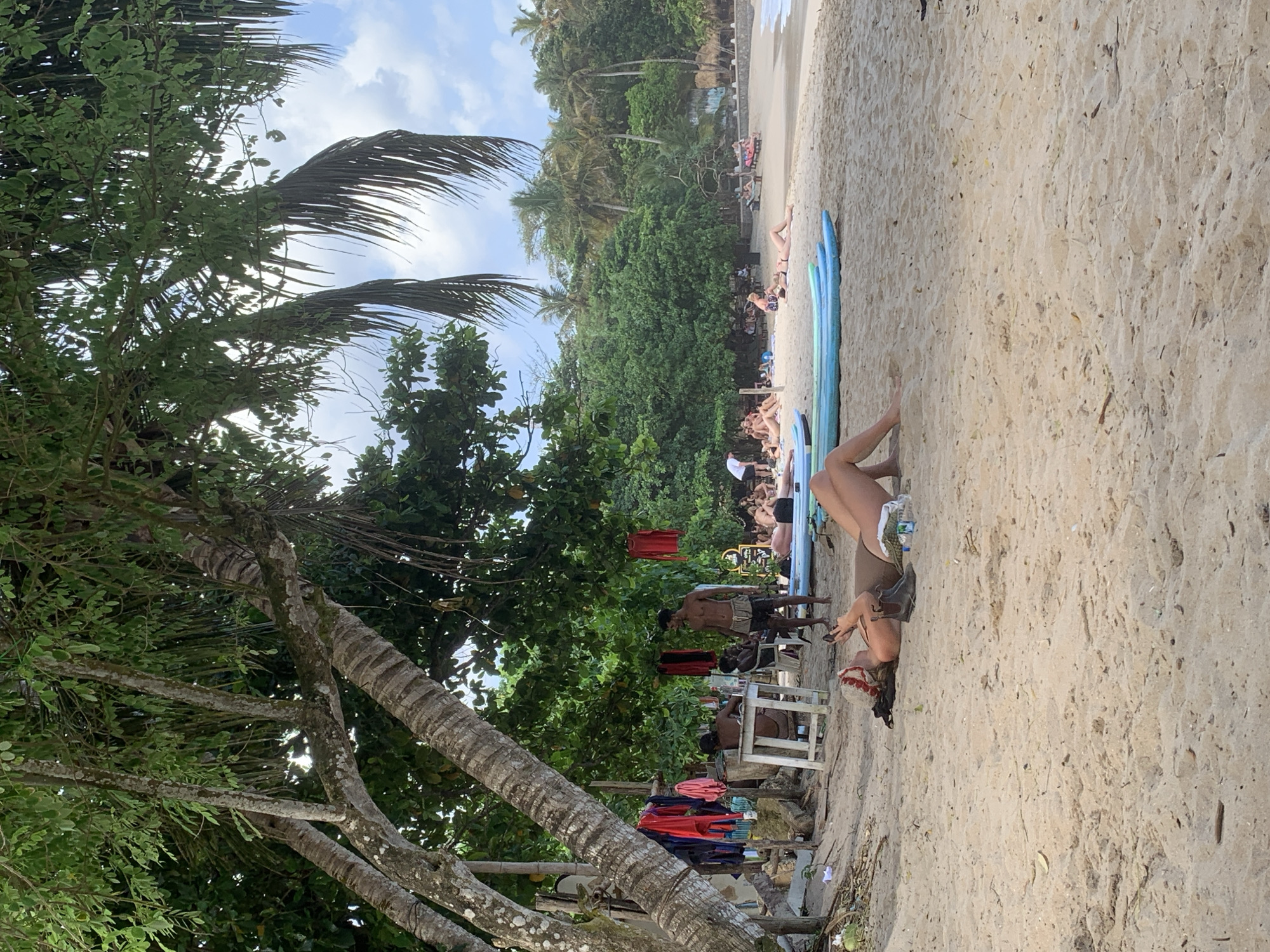 The image depicts a tropical beach scene in Dikwella, Sri Lanka, with a focus on the sandy shore, lush vegetation, and several people enjoying the environment. In the foreground, a person lies on their back on the sand, wearing a bikini, with a pair of dark boots and a bottle of water next to them. Further back, a row of surfboards rests on the sand, with several people sunbathing or relaxing on the beach. To the left, a makeshift stall or structure is visible, with clothes hanging on a line and a few people gathered around. A man stands near this stall, wearing shorts. Palm trees and other dense green foliage form a backdrop to the beach, with the ocean visible in the distance. The sky is partly cloudy. The overall impression is one of a relaxed, seaside atmosphere.