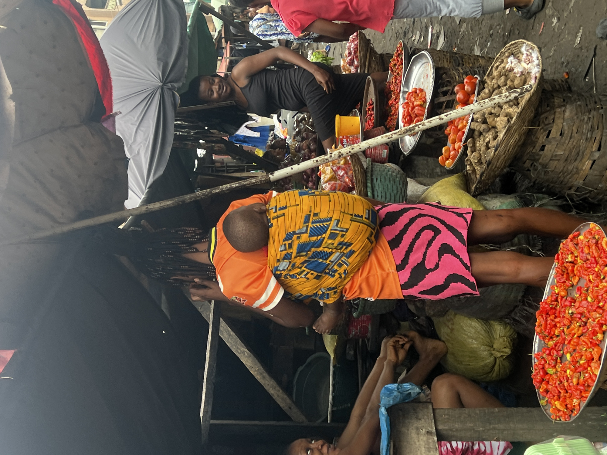 The image depicts a bustling outdoor market scene in Nkpor, Nigeria. A woman, from the rear, is carrying a child on her back in a vibrantly patterned yellow and blue wrap. She is wearing an orange t-shirt with white stripes on the sleeves and a brightly colored pink and black patterned skirt. To her right, another woman in a black sleeveless dress stands smiling and looking towards the left. Behind them, various market stalls are visible, laden with produce. In the foreground, a metal tray is filled with numerous small red peppers. Other baskets and trays contain tomatoes and what appear to be root vegetables. The market environment is implied by the presence of awnings, stalls, and the ground, which appears to be dirt or unpaved. The overall impression is one of lively commerce and daily life in a Nigerian market.