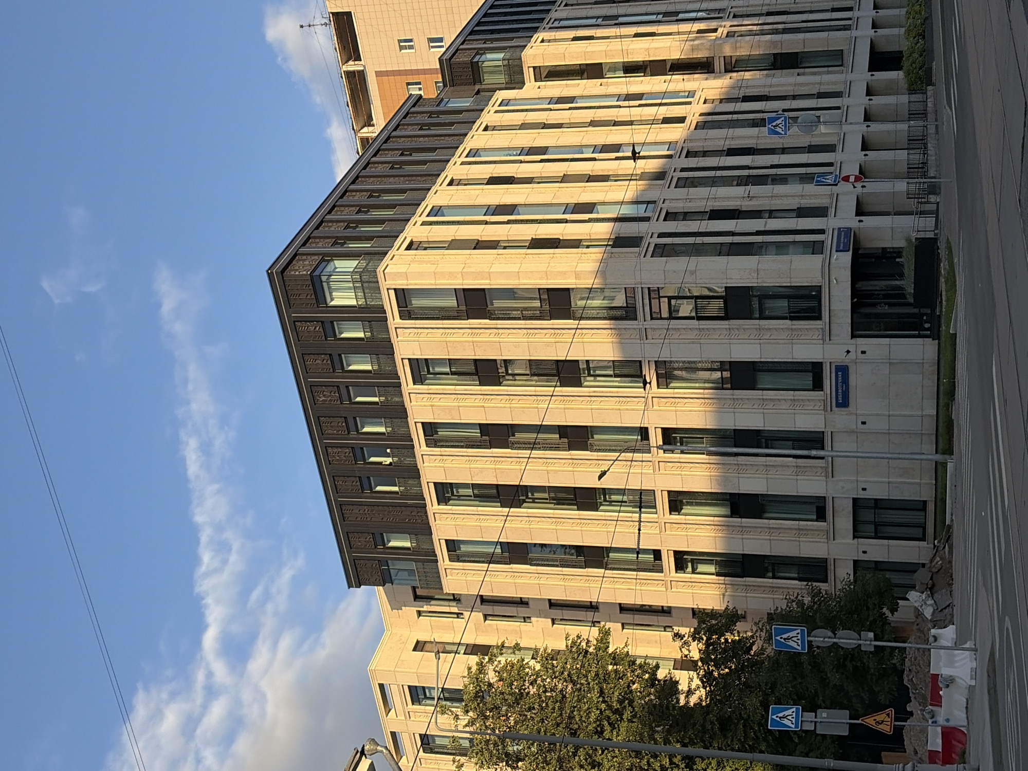The image shows a modern, light-colored stone building with dark-framed windows and balconies. The building has multiple stories and a dark, angular roofline. The sun is casting long shadows across the facade, indicating it is late afternoon or early morning. In the foreground, there is a street with crosswalk markings and some construction debris, including concrete barriers and piles of dirt. There are also traffic signs and lampposts visible. A green tree is partially visible on the left side of the image. The sky above is blue with scattered white clouds. A street sign in Russian, "Библиотечная улица" (Bibliotechnaya Street), is visible on the building. This suggests the location is Bibliotechnaya Street in Moscow, Russia. There are no people visible in the image.