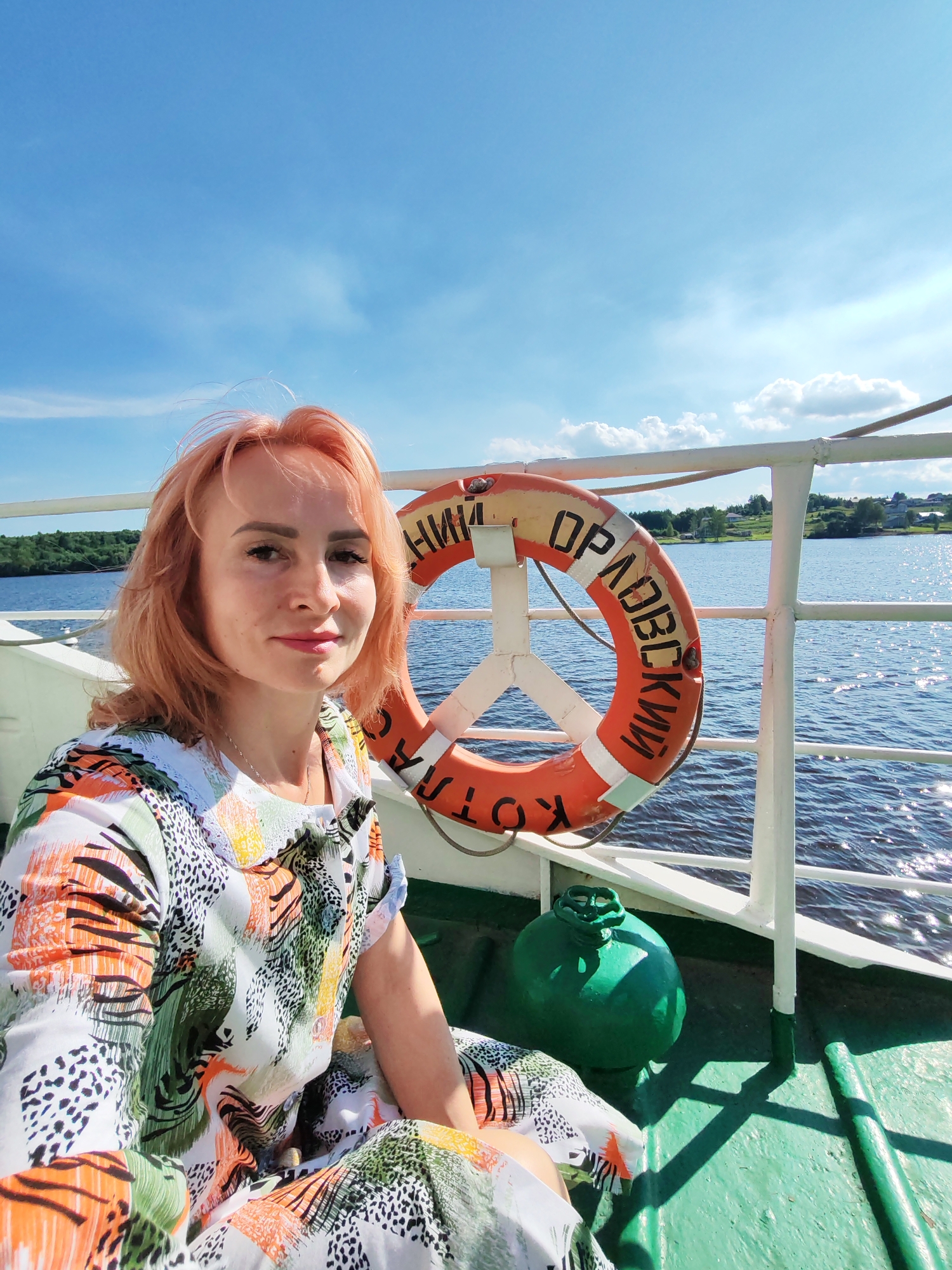 A woman with auburn hair is taking a selfie on a boat. She is wearing a colorful, patterned dress and is seated on the deck, looking at the camera with a slight smile. Behind her, a life preserver is hanging on a white railing. The life preserver is orange with white stripes and has Russian text on it that appears to read "КОТЛОВСКИЙ". The boat is moving on a body of water, and the sky is bright blue with scattered clouds, suggesting it is daytime and the weather is fair. The background shows a green, tree-lined shore with some buildings in the distance. A large green object, possibly a buoy or a piece of equipment, is visible on the deck next to the woman. The overall scene conveys a sense of leisure and travel.