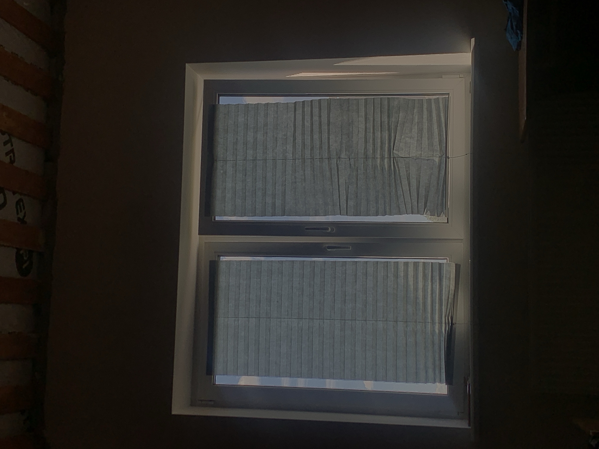 The image shows a white window frame with two panes, each covered by grey pleated blinds. The blinds are partially lowered, allowing some light to filter through. The window is set into a dark wall, and the top of the frame is near the ceiling, which has exposed wooden beams and insulation. A portion of a shelf with a blue cloth on it is visible to the right of the window. The scene appears to be indoors, likely in a room under construction or renovation, given the exposed ceiling elements. There are no people or animals present in the image. The lighting suggests it is daytime, with sunlight entering from the left side of the window. The image does not contain any specific landmarks or details that would allow for pinpointing a location within Minsk, Belarus.
