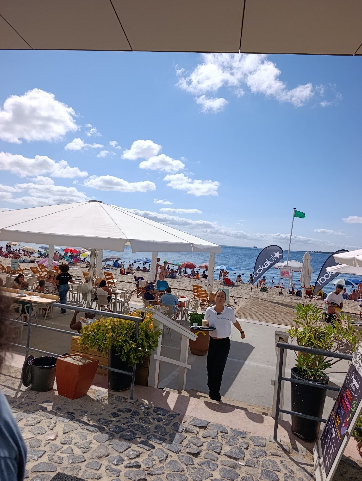 The image depicts a sunny day at a beachside establishment in Setúbal, Portugal. In the foreground, a woman wearing a white crop top and black pants, who appears to be a server, walks up a set of stairs holding a tray with drinks. Behind her, a black trash can and an orange planter are visible. The background reveals a crowded beach with people sunbathing and swimming in the ocean. Large white umbrellas provide shade for tables and chairs set up on the sand, suggesting an outdoor dining area. Several flags are planted on the beach, including a green flag and two flags with the logo "rockalot". The overall scene conveys a lively atmosphere at a popular beach destination.