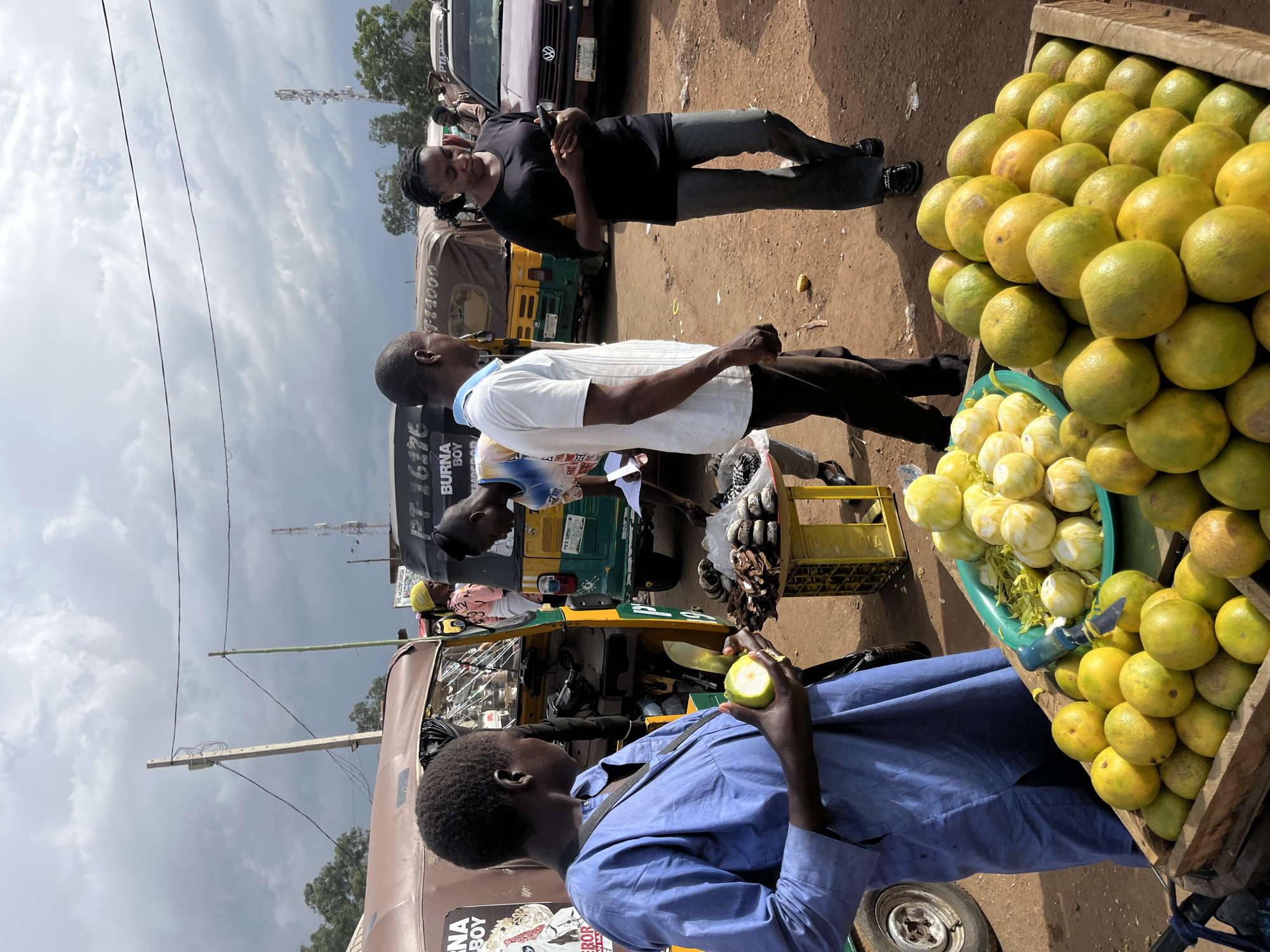 The image shows a bustling outdoor market scene, likely in Rafin Par, Nigeria. Three individuals are prominently featured, with a focus on their interactions and surroundings.
In the foreground, a person wearing a blue collared shirt is holding a green fruit, possibly a lime or a small unripe orange, and examining it. Their attention is directed towards the fruit, suggesting a purchase or inspection. Behind them, a wooden crate overflows with a variety of citrus fruits, primarily oranges and lemons, with some greener fruits mixed in. A green plastic basin filled with lemons is also visible.
Further back, another person in a white collared shirt with a blue collar is engaged in conversation with a third individual wearing a black t-shirt. They appear to be standing near a three-wheeled vehicle, possibly a Bajaj auto-rickshaw, which is common in many parts of Africa. The vehicle has a brown cover and some signage on its side, though the text is partially obscured and difficult to read. The overall atmosphere suggests a lively marketplace with vendors and customers.
The sky is cloudy, indicating potentially overcast weather. The ground is made of dirt, characteristic of many rural or semi-urban market settings. The time of day appears to be daytime, based on the lighting conditions. The presence of the auto-rickshaw and the types of fruits suggest a tropical or subtropical region.
There is visible text on the side of the auto-rickshaw, including "BURNBOY," which could be a brand or a reference to a person. Another partially visible text on the vehicle reads "PT-16," possibly a license plate number or identification.
The image captures a moment of everyday life in a Nigerian market, highlighting the interaction between people and the goods they are selling or buying.