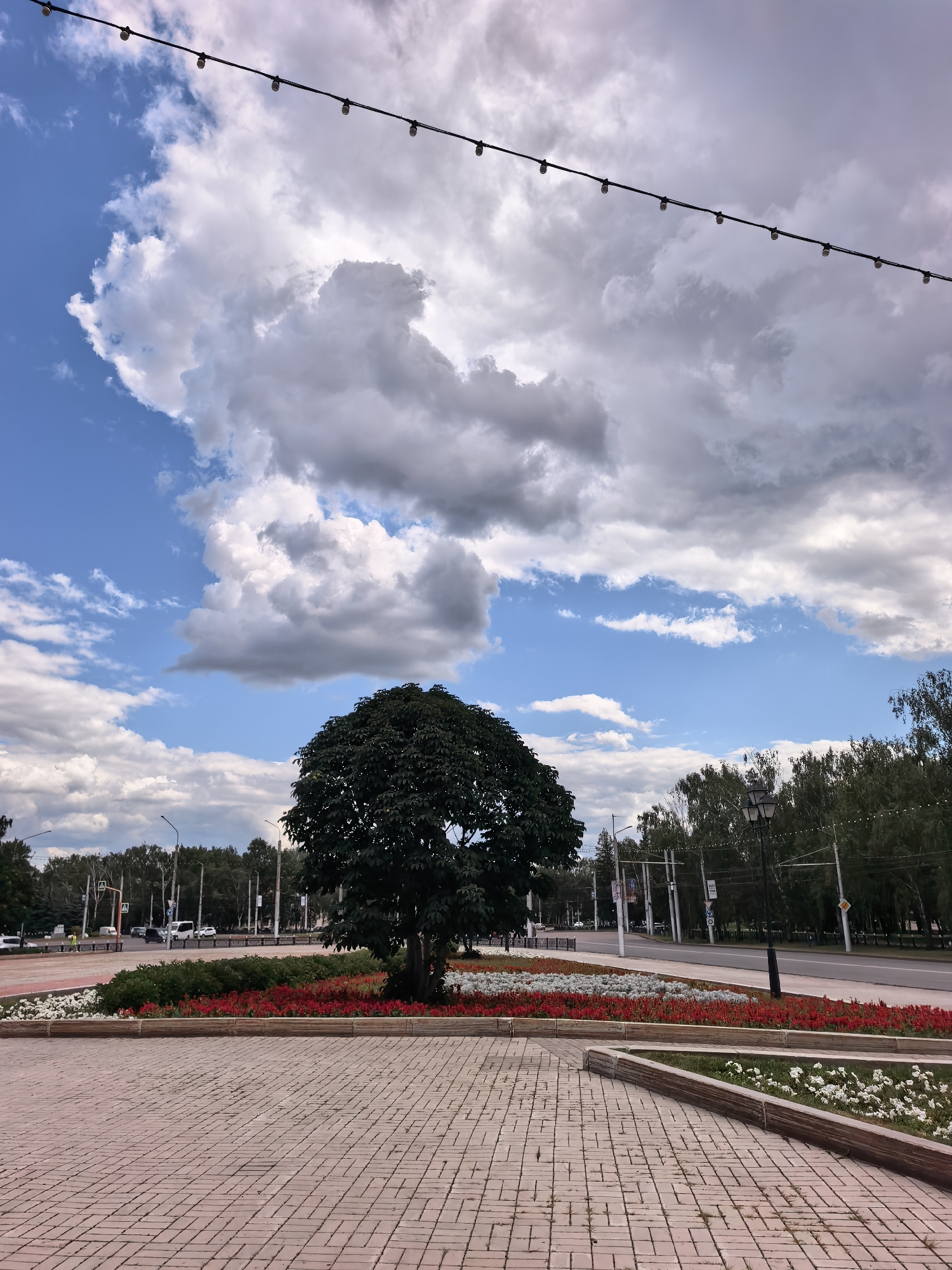 This is a photograph of a street scene in Sterlitamak, Russia. The foreground is a paved area with a reddish-brown brick pattern. In the middle ground, there is a landscaped island with a large, dark green tree at its center. This island is bordered by beds of red and white flowers. Beyond the island is a road with cars and traffic lights. A line of trees runs along the far side of the road, and in the distance, there are buildings. The sky is blue with large, puffy white and gray clouds. A string of small, bare lightbulbs is visible across the top of the image. There are no people clearly visible in the photograph.