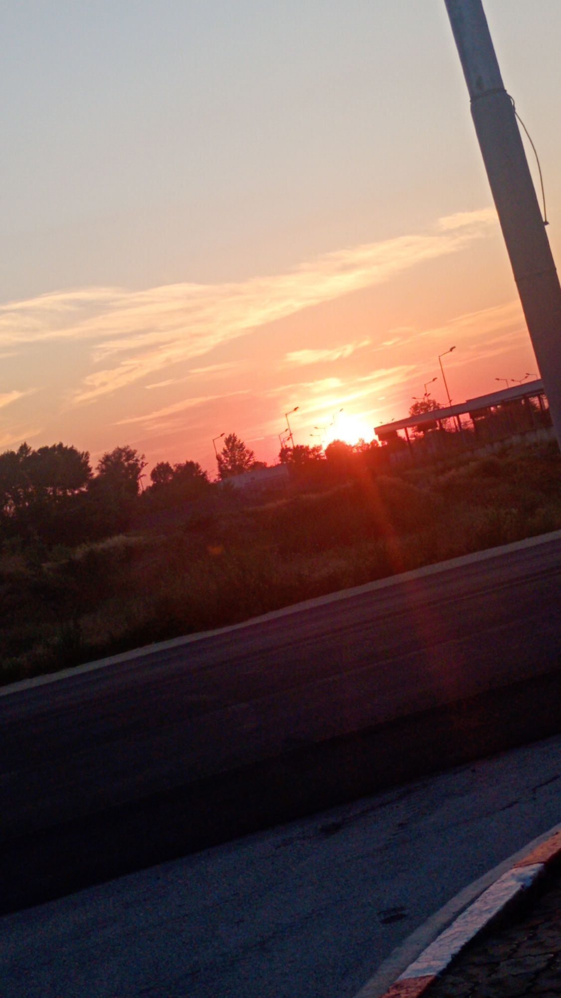 A vertical, slightly tilted photograph captures a sunset over Aidemir, Bulgaria. The sky is a gradient of pale blue at the top transitioning to vibrant orange and pink near the horizon. Wispy clouds are scattered across the sky, catching the sunset's light. The sun itself is low on the horizon, appearing as a bright, slightly diffused orb with visible sun rays.
In the middle ground, a line of trees and vegetation creates a dark silhouette against the bright sky. Several streetlights are visible among the trees, some illuminated, others not. A long, low structure, possibly a building or a covered walkway, is situated behind the trees.
The foreground is dominated by an asphalt surface, which appears to be a road or pavement. The surface is angled diagonally from the bottom left corner upwards towards the right. A white line marking is visible on the right side of the asphalt, indicating an edge or lane divider. In the far right of the image, a tall, dark pole, possibly a lamppost or utility pole, extends vertically, partially obscuring the sunset. The overall impression is that of an outdoor scene at dusk, possibly near a road or public space.
