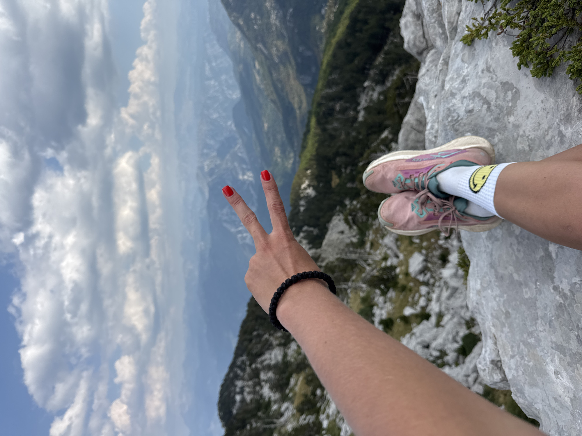 In the city of Mrtvo Duboko, Montenegro, a person sits on a rocky outcrop, their legs crossed. They are wearing pink athletic shoes and white socks adorned with a yellow smiley face. A black beaded bracelet is on their left wrist, and their hand is raised, displaying a peace sign. The background is a dramatic mountainous landscape with layered peaks fading into the distance under a cloudy sky. The scene is captured from a first-person perspective, focusing on the person's limbs and the expansive natural scenery.