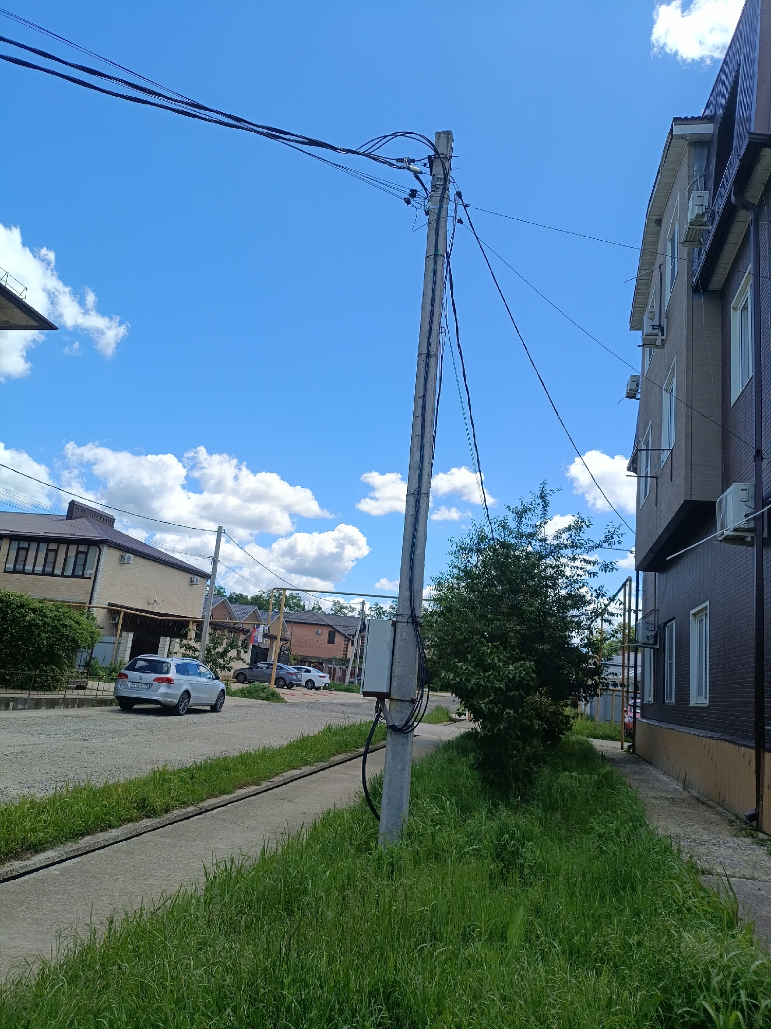 The image captures a residential street scene on a bright, sunny day, likely in Goryachiy Klyuch, Russia. In the immediate foreground, a tall, grey concrete utility pole dominates the center, festooned with numerous black electrical and communication wires that crisscross the sky and connect to the buildings. A grey electrical box is mounted on the pole. Overgrown green grass flanks a narrow concrete path or road that runs along the right side of the frame and appears to be part of a larger unpaved road to the left.
In the midground, to the left, a silver Volkswagen Passat station wagon with the license plate `P612YY 123` is either parked or slowly moving on a dirt road. Behind it, a two-story house with light-colored walls and a dark roof is visible, along with other residential buildings further down the street. To the right, a darker-colored, two-story building with multiple outdoor air conditioning units affixed to its exterior and white-framed windows stands close to the path. A lush, bushy green tree is situated between the utility pole and this building.
The background features a clear blue sky dotted with scattered white cumulus clouds, suggesting fair weather. No people are visible, and there is no explicit activity taking place beyond the static presence of buildings, vehicles, and natural elements. The scene conveys a quiet, suburban atmosphere.