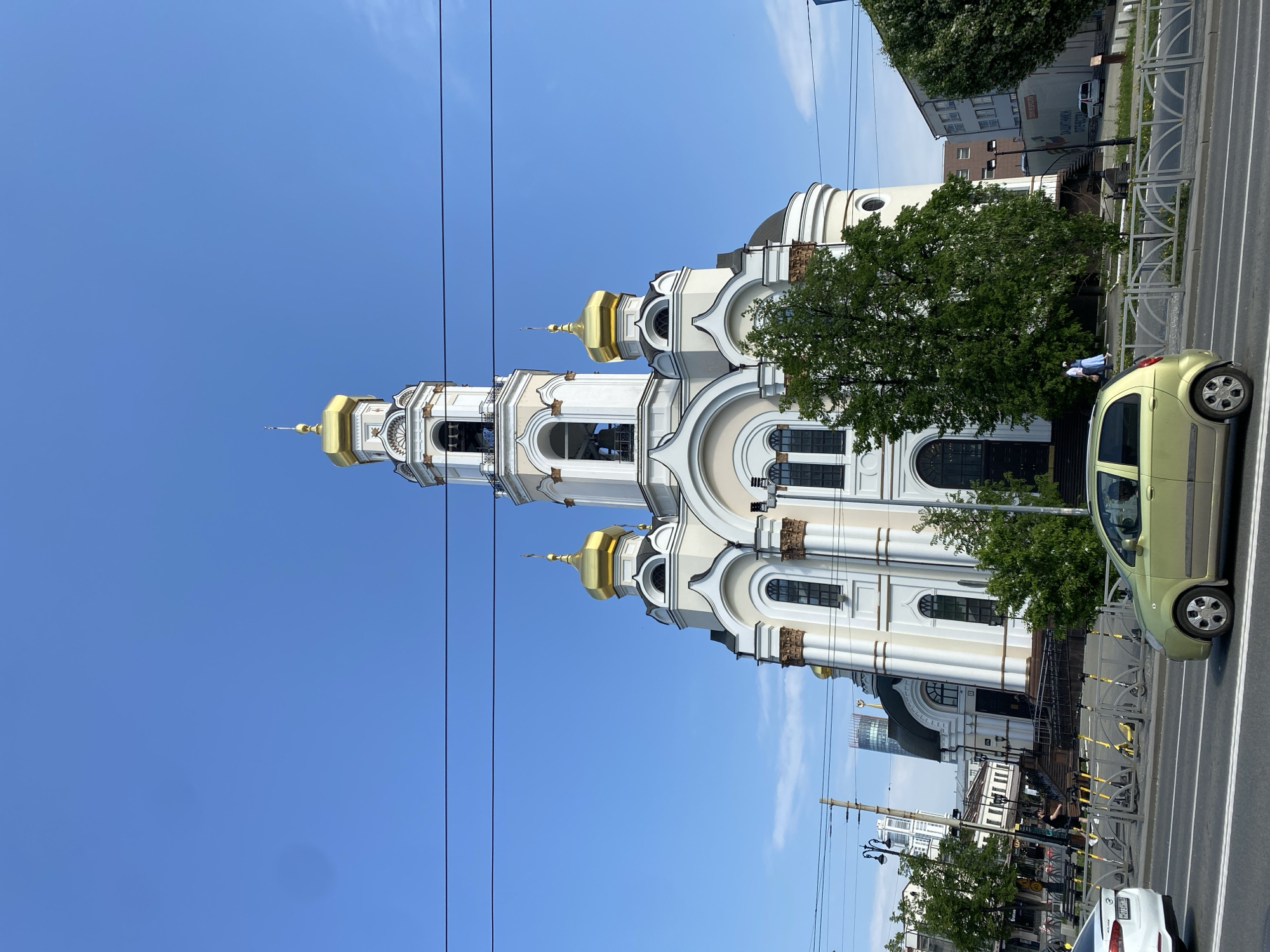 The image depicts the Church on Blood in Yekaterinburg, Russia. The church, with its white walls and golden domes, stands tall against a clear blue sky. Power lines are visible in the foreground. A lime green car is parked on the street beside the church. Trees partially obscure the view of the building. Pedestrians are present on the sidewalk near the church. The image appears to be taken from street level.
