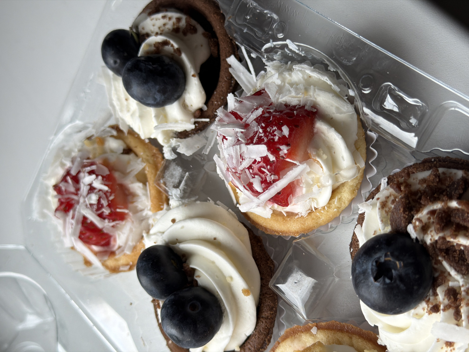 A high-angle, close-up shot captures a selection of miniature tarts or pastries nestled within a clear plastic container. The scene is static, focusing entirely on the confectionery.
The container holds at least six small desserts, arranged in molded compartments. The pastries feature two main varieties:
1. **Dark Crust Tarts:** These have a dark, possibly chocolate-flavored, crust. They are topped with generous swirls of white whipped cream or frosting and adorned with two whole, dark blueberries. Some of these tarts also have sprinkles of dark chocolate shavings on the cream.
2. **Light Crust Tarts:** These feature a lighter-colored, possibly shortbread or vanilla, crust. They are also topped with white whipped cream or frosting, over which a bright red, glossy strawberry topping or compote is spread. These are generously sprinkled with delicate white chocolate shavings.
The plastic container is clear and shows reflections, indicating a smooth, shiny surface. The edges of the container's lid are visible at the top and right, suggesting it's either partially open or a close-up of the tray. The background is a plain, light surface, likely a table.
There is no activity taking place, as it's a still life of prepared food. No people are visible, and no text is discernible on the packaging or pastries. The image provides no direct visual cues for time of day or specific location, though it is contextualized as being from Penza, Russia. The focus is on the appealing presentation of the sweet treats.