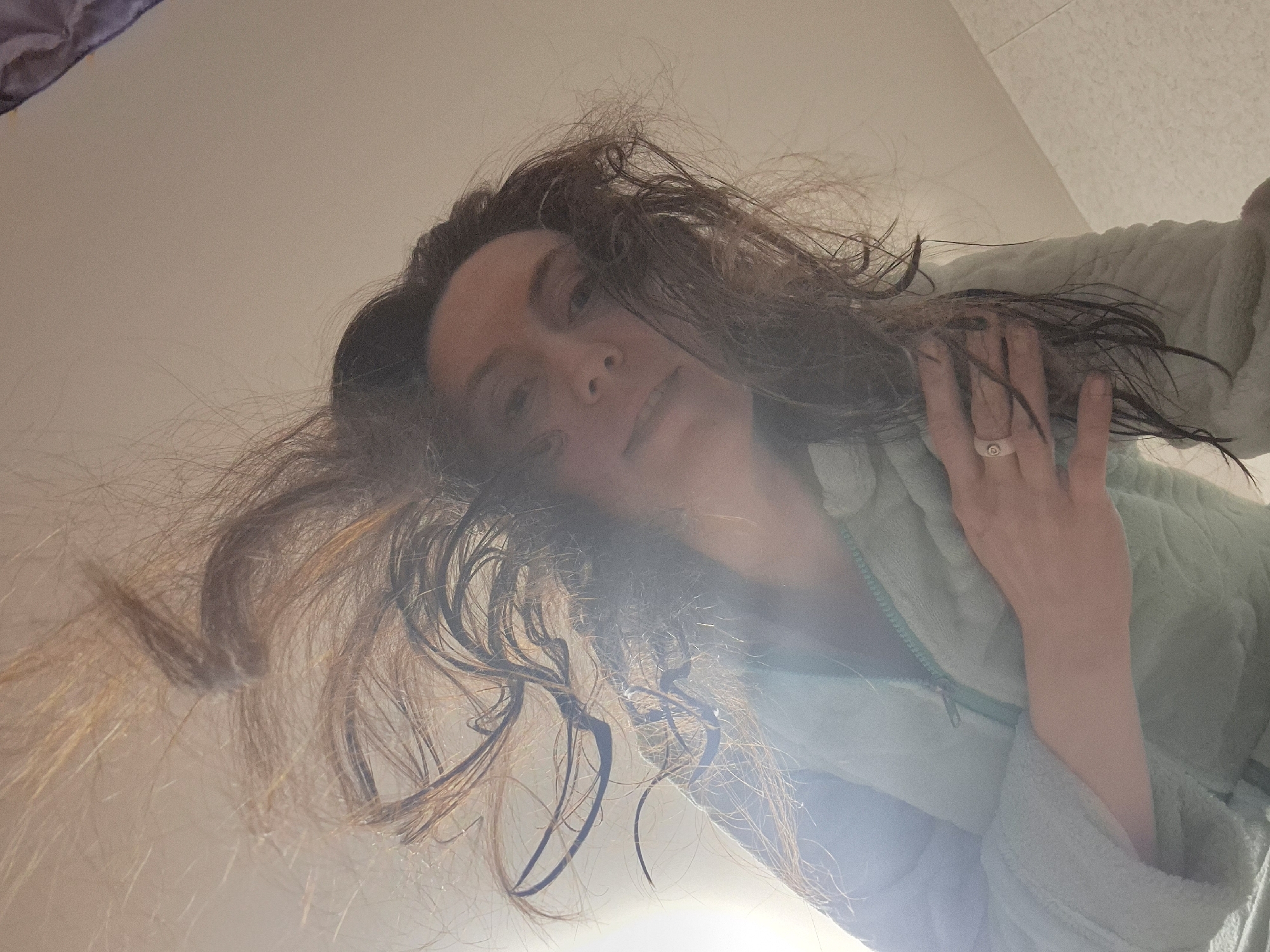A low-angle shot depicts a person with long, wet brown hair and a light complexion, wearing a light green fuzzy bathrobe. The person's face is slightly out of focus and positioned towards the upper center of the frame. Their right hand, adorned with a ring, is raised and partially obscures the right side of their face. The background consists of plain white walls, with a hint of purple visible in the upper left corner. A bright light source illuminates the scene from below, causing glare and contributing to the overall soft focus. The location is Unknown, Unknown.