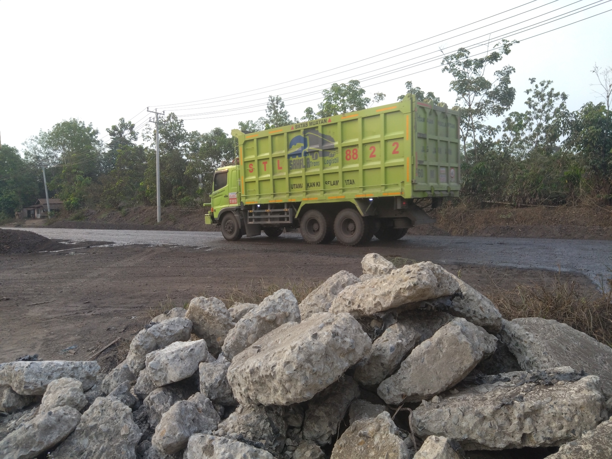 A light green dump truck, identified by the markings "STL SAHILA TRANS Logistik" and the numbers "88 2" on its side, is parked on a dirt road. The truck's bed is empty. In the foreground, a pile of broken concrete debris lies on the ground. The background features a line of trees and power lines stretching across a bright, overcast sky. The scene appears to be in Sedupi, Indonesia.