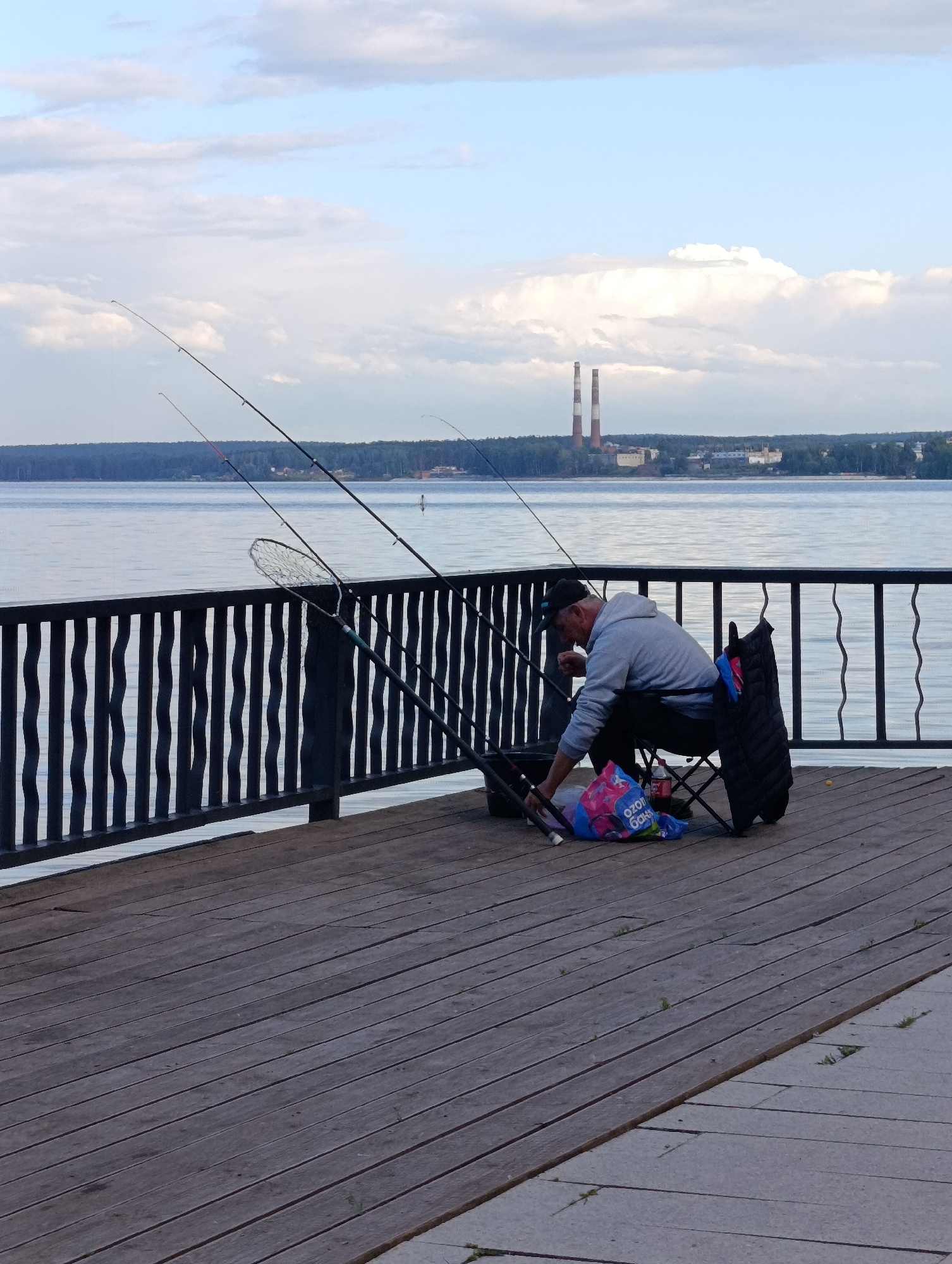 A man is fishing on a wooden pier in Chaikovsky, Russia. He is sitting on a folding chair with a gray hoodie and black pants. A fishing net is propped up next to him, and several fishing rods are in the water. In the background, there is a body of water, a forested shoreline, and two large smokestacks with red and white stripes. The sky is cloudy.