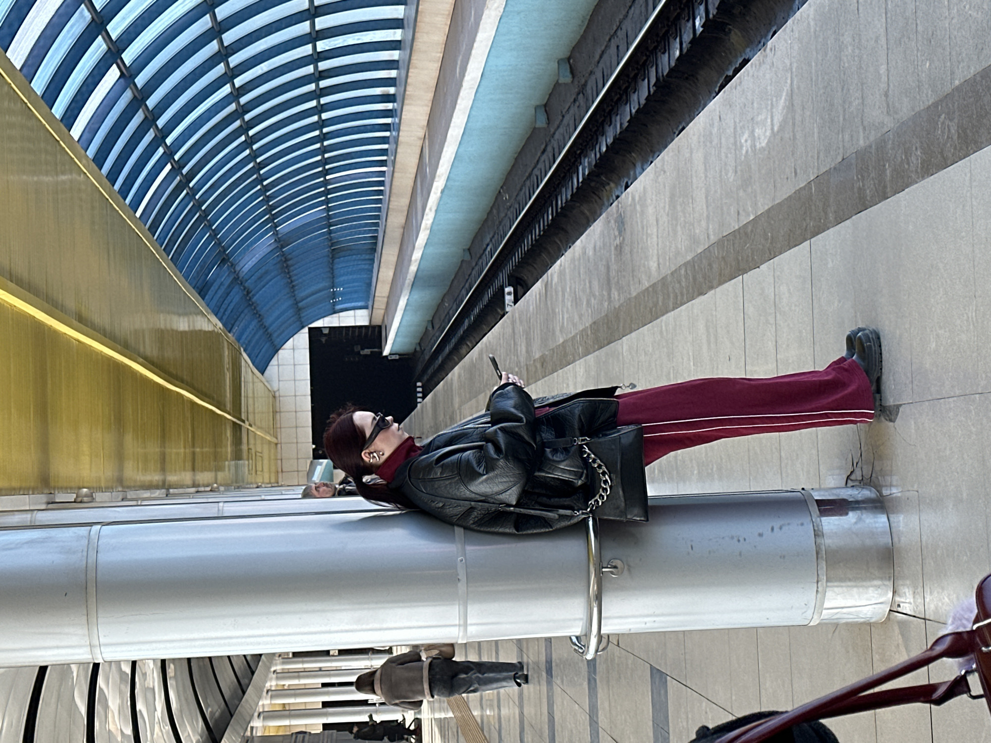 An outdoor metro station in Kazan, Russia, is depicted from a low-angle perspective. The main subject is a female with reddish-brown hair tied back, wearing dark sunglasses, a black turtleneck, an oversized black leather jacket, and dark red track pants with white vertical stripes. She is leaning against a large, cylindrical, light-grey metal pillar on the platform, facing downwards and actively interacting with a black smartphone held in both hands. A small black shoulder bag with a silver chain strap hangs from her left shoulder. Multiple silver-toned earrings are visible on her left ear.
The platform floor is composed of light grey tiles with dark grey demarcation lines. To the right, a single train track is visible, bordered by a light blue wall and a light brown ledge. The track appears damp in sections. The station ceiling features a distinctive design: a vaulted, blue and white striped arched section extends over the tracks, while a reflective, golden-yellow panel covers the area above the platform. In the background to the left, another individual is seen from behind, walking away, wearing a light brown jacket and dark trousers, carrying a bag. A partial view of a male's head and shoulder is visible behind the central pillar. A dark red handbag is partially visible in the bottom left foreground. The scene suggests a typical moment of waiting at a transit hub.