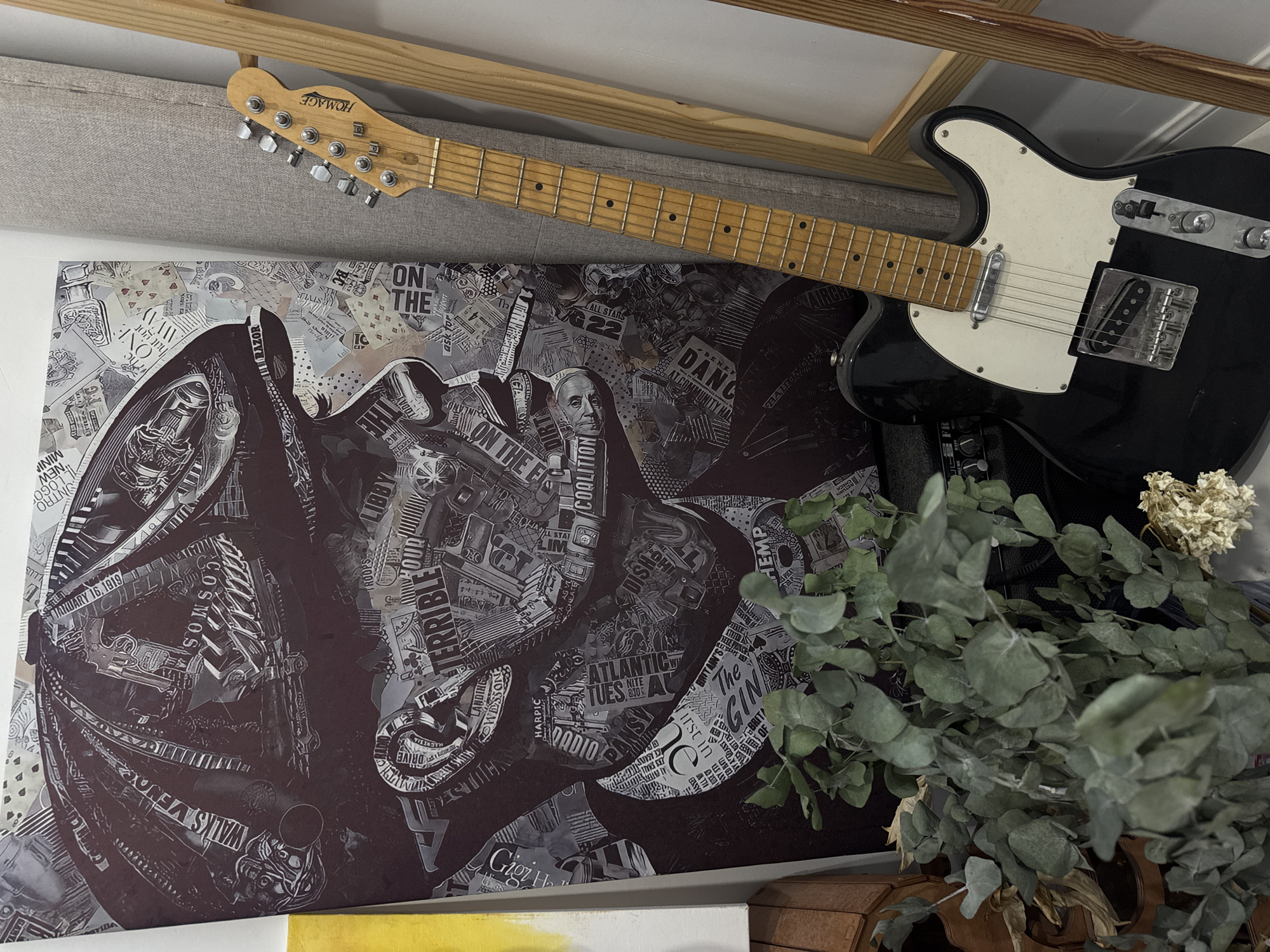 The image depicts an indoor scene, likely a room, featuring a large, dark-toned collage artwork on a canvas. The artwork is abstract and appears to be composed of various printed materials. Leaning against the artwork is a black electric guitar with a light-colored neck and a white pickguard. To the right of the guitar is a light-colored wooden ladder leaning against a textured, gray fabric surface. In the foreground, a bundle of dried eucalyptus branches with green leaves and some dried flowers are placed in front of a black amplifier. The overall lighting is dim, suggesting an indoor setting without direct sunlight. There are no people visible in the image. The location is not specified as Oral, Kazakhstan, nor is there any indication within the image itself to suggest this geographical context.