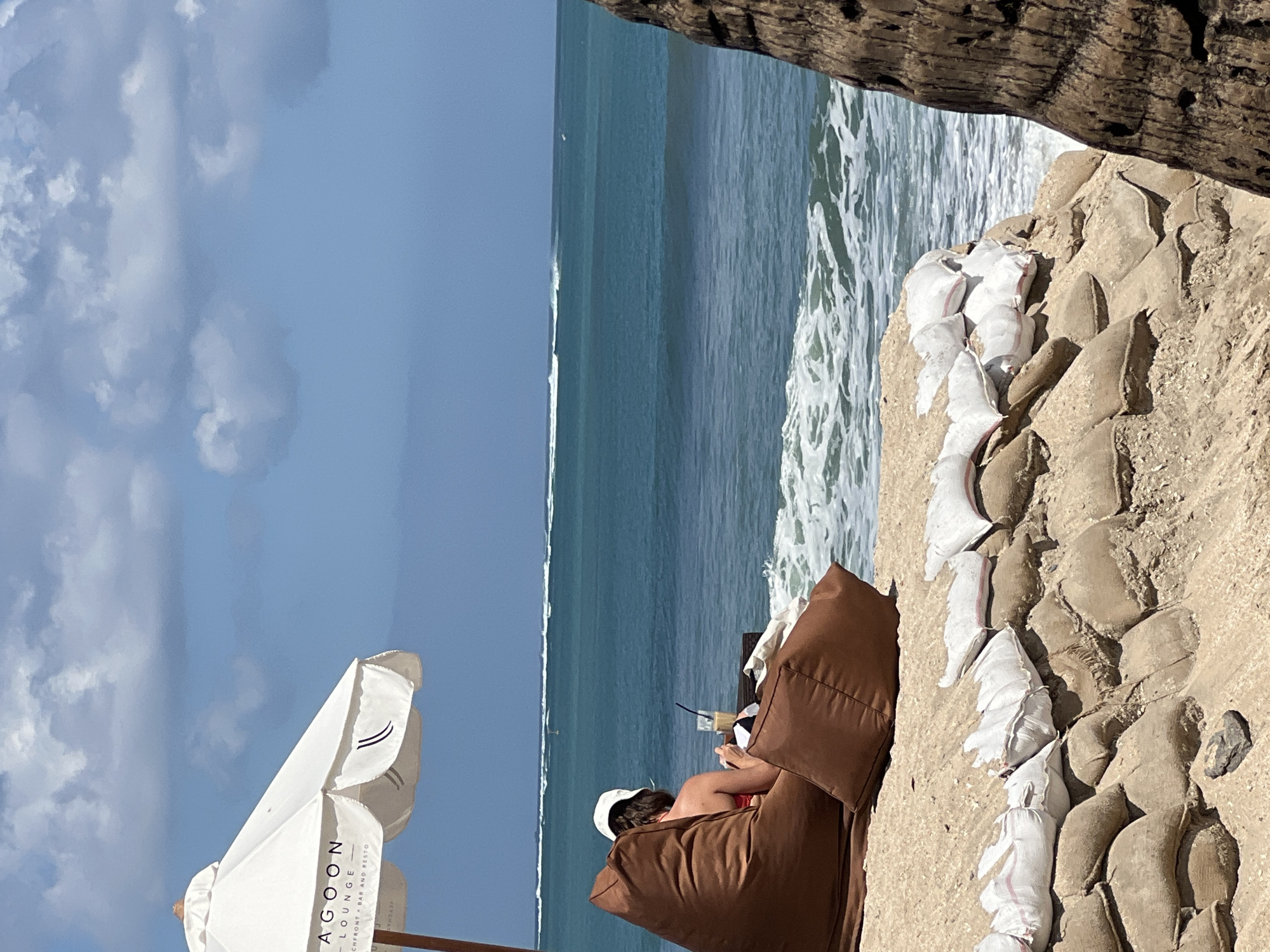 A person sits on a brown beanbag chair on a sandy beach, facing the ocean. The person is wearing a white hat and a red swimsuit. In front of the person is a table with a drink. A white umbrella with the text "LAGOON LOUNGE" is to the left of the person. The ocean has small waves, and there are sandbags stacked along the edge of the beach, presumably to prevent erosion. The sky is blue with white clouds. The scene suggests a relaxed atmosphere at a beachfront establishment in Kuta, Indonesia.