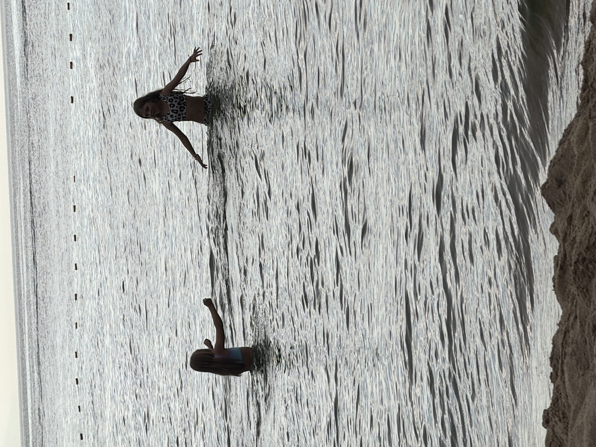 This media file shows two young girls playing in the water at a beach. The image is captured from a high angle, looking down at the water. The water is choppy and reflective, with sunlight glinting off the surface. The sand on the right side of the image appears dry and coarse.
In the upper part of the image, one girl is visible from the waist up, wearing a leopard-print bikini. She is looking towards the camera with a slight smile and her arms are outstretched, as if she is posing or playing. Her hair is long and dark, and it appears wet.
In the lower part of the image, another girl is visible from the shoulders up, wearing a patterned swimsuit. She is facing away from the camera, with her head tilted to the side. Her right arm is raised with her fist clenched.
The scene appears to be during the daytime, with bright sunlight. The overall mood is playful and joyful, suggesting a summer day at the beach. There is no visible text in the image. Given the location context of Goryachinsk, Russia, this is likely a scene at Lake Baikal, known for its sandy beaches and clear waters. The vertical orientation of the image suggests it was taken with a phone held in portrait mode.