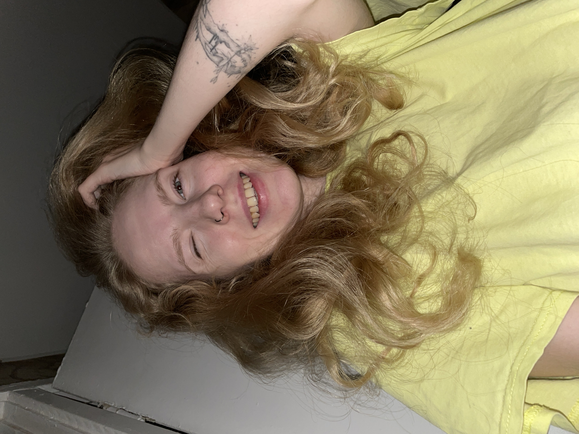 A woman with long, wavy red hair lies on her back, wearing a yellow shirt. Her right arm is raised, with her hand in her hair. She is smiling and winking. A tattoo is visible on her right arm. The background consists of a white surface and a dark area. There is no information in the image to suggest that it was taken in Izhevsk, Russia.