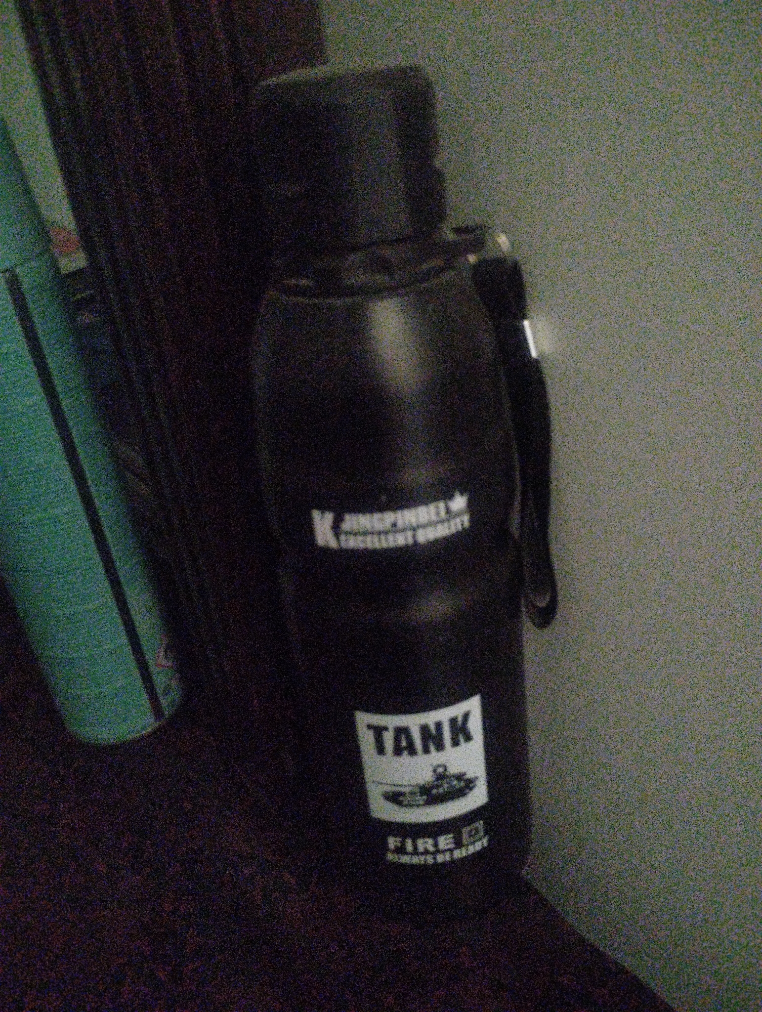 A dark, grainy image shows a black thermos bottle with white lettering. The bottle has a handle and a cap. On the side of the bottle, the word "TANK" is printed in large white letters, with a depiction of a tank below it. Further down, the word "FIRE" is also visible in white letters. To the left of the bottle, a portion of a green object is visible, possibly a book or a rolled-up item. The background is dark and out of focus, with a textured wall on the right side of the image. The lighting is dim, and there is significant noise in the image.