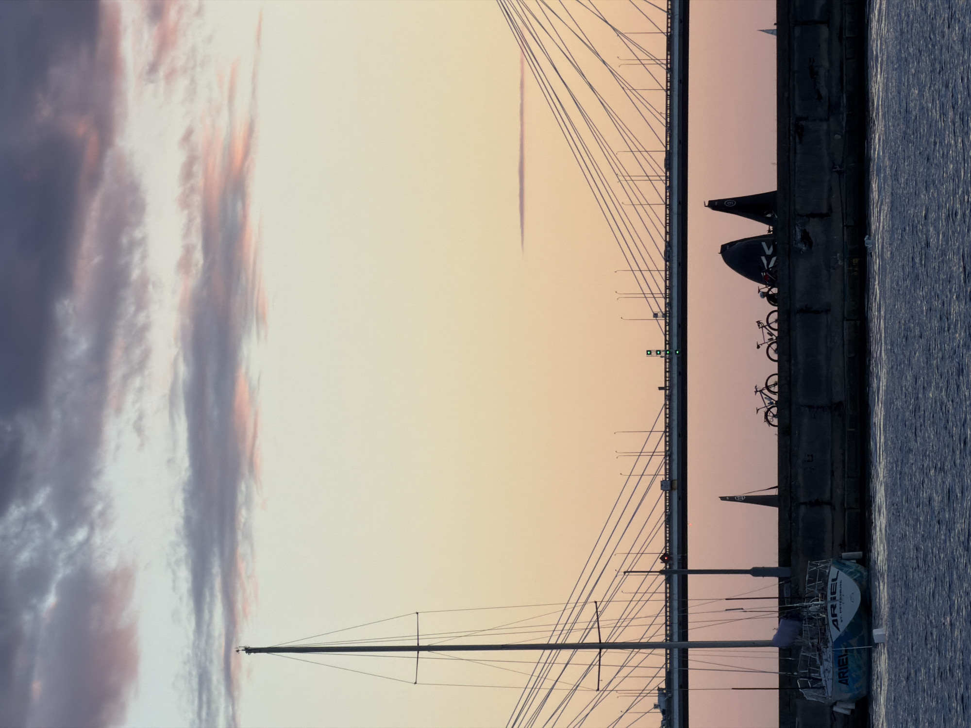 The image depicts a view of the Neva River in Saint Petersburg, Russia, at dusk. A large cable-stayed bridge spans the river in the background, with vehicles visible crossing it. In the foreground, a sailboat named "ARIEL" is moored to a dock. The sailboat has the inscription "ST. PETERSBURG" on its hull. A concrete embankment runs along the river, on top of which are several bicycles and two dark, sail-like structures. The sky is filled with soft, pastel clouds tinged with pink and purple hues. The water in the river is dark and calm. The overall scene conveys a sense of tranquility and urban serenity as day transitions into night.