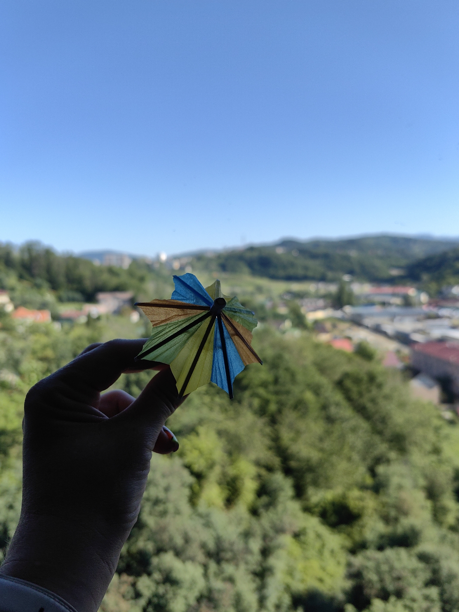 A hand holds a colorful, miniature cocktail umbrella against a blurred, green, hilly landscape under a clear blue sky. The umbrella has alternating panels of blue, yellow, and brown. The hand is silhouetted, with the exception of some fingernails painted with red and green polish. In the background, a town or city is visible with red-tiled roofs and buildings scattered amongst dense green foliage and rolling hills, suggestive of the mountainous terrain surrounding Sochi, Russia.