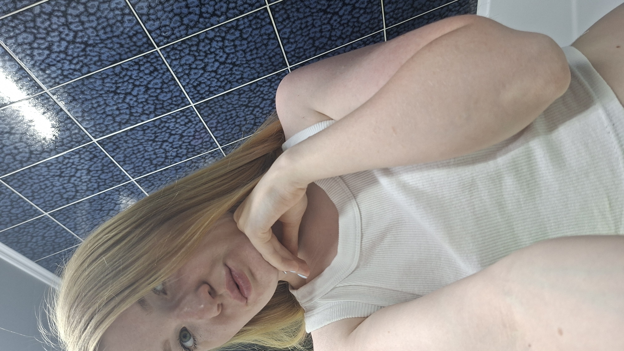 A fair-skinned person with blonde hair is lying on their back with a blue patterned tile floor behind them. The person is wearing a white ribbed tank top and their right hand is touching their face. The location is not discernible as Chaikovsky, Russia.