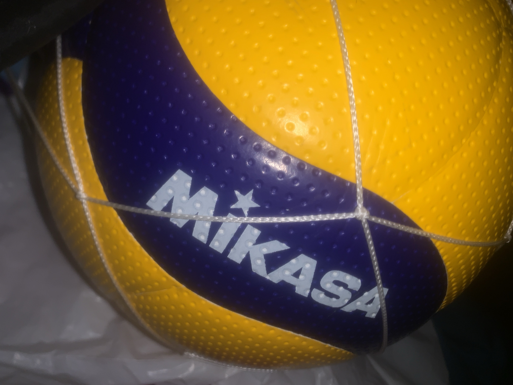 The image displays a close-up shot of a Mikasa volleyball, partially enclosed within a white, coarse-mesh net. The ball is predominantly yellow with a large, curved blue section. Both the yellow and blue areas feature a distinct textured, dimpled surface. The brand name "MIKASA" is clearly visible in white capital letters on the blue segment, with a white star design positioned above the 'K' and 'A'. The volleyball rests on a crumpled white plastic material, possibly a bag or sheeting. The background is dark and indistinct, suggesting an indoor setting or an area with low lighting. The white net wraps around the ball, with its strands visible across both the yellow and blue portions. No activity or people are depicted; the focus is solely on the volleyball. This image was taken in Vsevolozhsk, Russia.