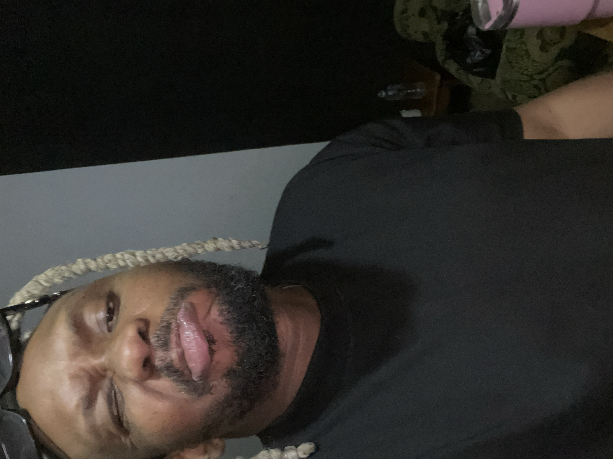 The image features a dark-skinned man with a beard and light-colored, rope-like braided hair, wearing glasses pushed up onto his head. He is captured in a close-up, rotated view, appearing to lie down or lean back. His expression is playful, with his mouth open and tongue slightly out, and one eye appearing to be closed in a wink or squint. He is wearing a plain black t-shirt.
The setting appears to be indoors with dim lighting, suggesting evening or night. The background consists of a dark upper section, possibly a wall, and a lighter grey surface below, likely a bed or couch. In the upper right background, a pink tumbler or cup is visible, along with a small clear bottle and a dark, patterned fabric, possibly part of a blanket or furniture. The overall mood conveyed by the man's expression is relaxed and humorous. The activity is a casual pose for a photograph, possibly a selfie. Given the context, this scene takes place in Jos, Nigeria. No visible text is present in the image.