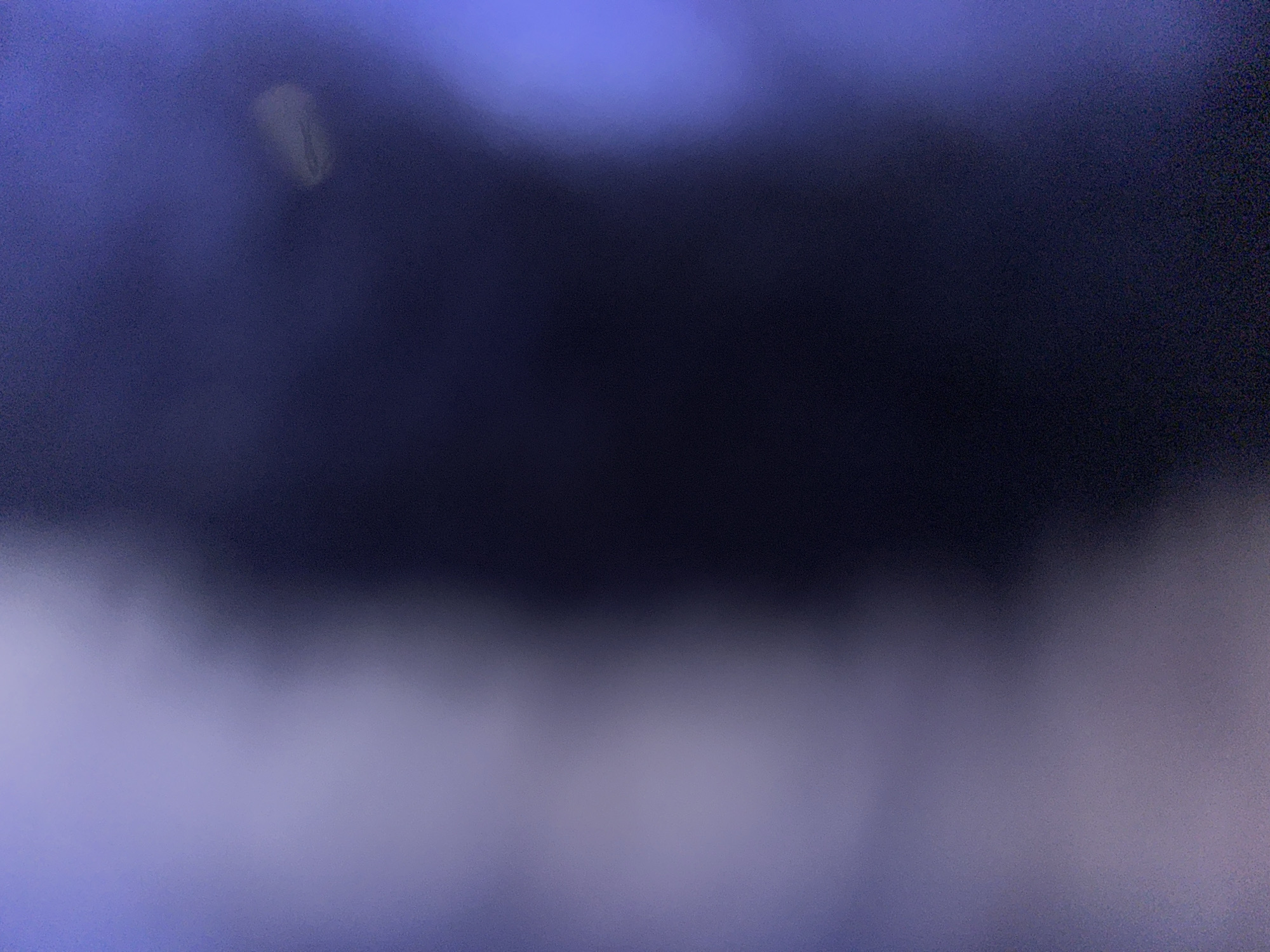 This is an abstract, out-of-focus image of a dark, possibly dimly lit environment. The dominant colors are dark blues and purples, with a lighter, hazy area on the left side of the frame. There are some indistinct bright spots, one of which appears as an oval shape in the upper right quadrant, suggesting light sources or reflections. The overall impression is of a blurry, atmospheric scene with no discernible objects or figures.