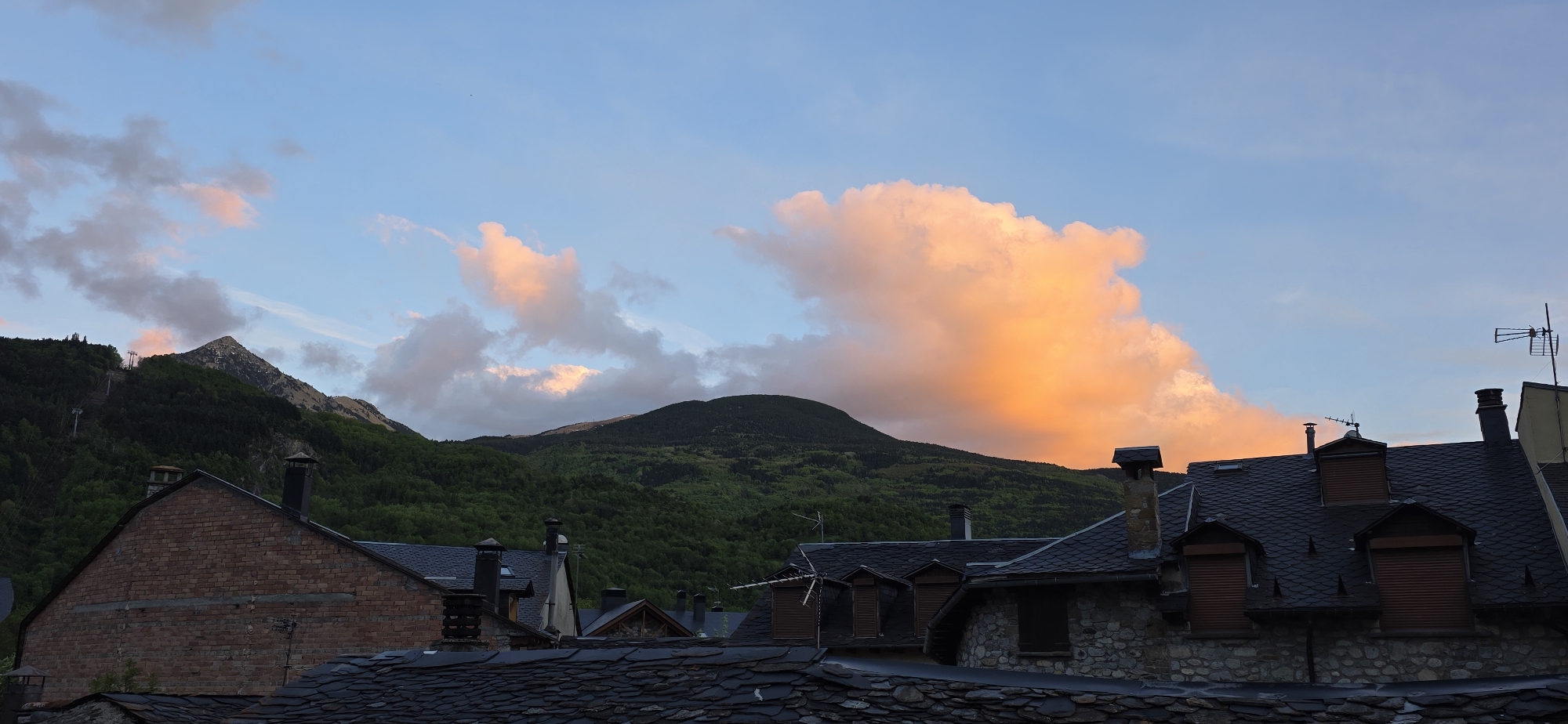 The image captures a scene in Benasque, Spain, showing a combination of natural landscape and architecture under a partially cloudy sky. The foreground is dominated by rooftops covered with dark grey, overlapping slate tiles. These rooftops are part of stone and brick buildings, some of which have chimneys and dormer windows with closed brown shutters.
Behind the buildings is a range of green mountains with a prominent, pointed peak visible on the left. The sky is light blue with clouds that are tinted with orange hues from the setting or rising sun. The clouds range from smaller, scattered formations to a larger, more substantial cloud mass on the right side of the image. There is an antenna on a rooftop on the right. The overall impression is a landscape where the built environment is integrated with the natural surroundings.