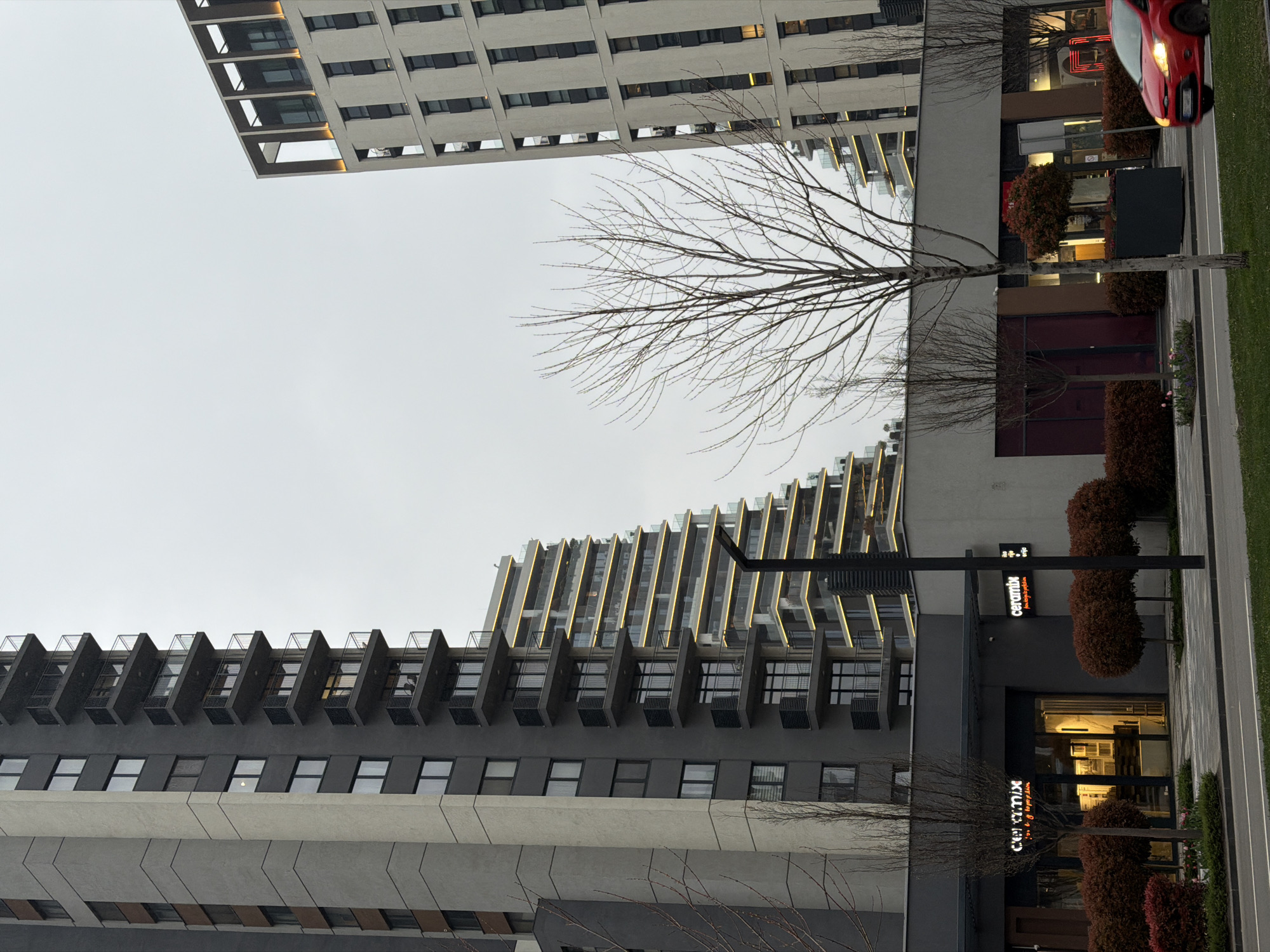 An eye-level outdoor shot depicts buildings and streetscape in Belgrade, Serbia. The sky is overcast. In the foreground are tall modern buildings, one gray and one white. Each building has a repetitive window pattern. In front of the gray building is a streetscape with trimmed bushes, a bare tree, and a sign for "ceramix". A red car is parked on the street next to the white building.