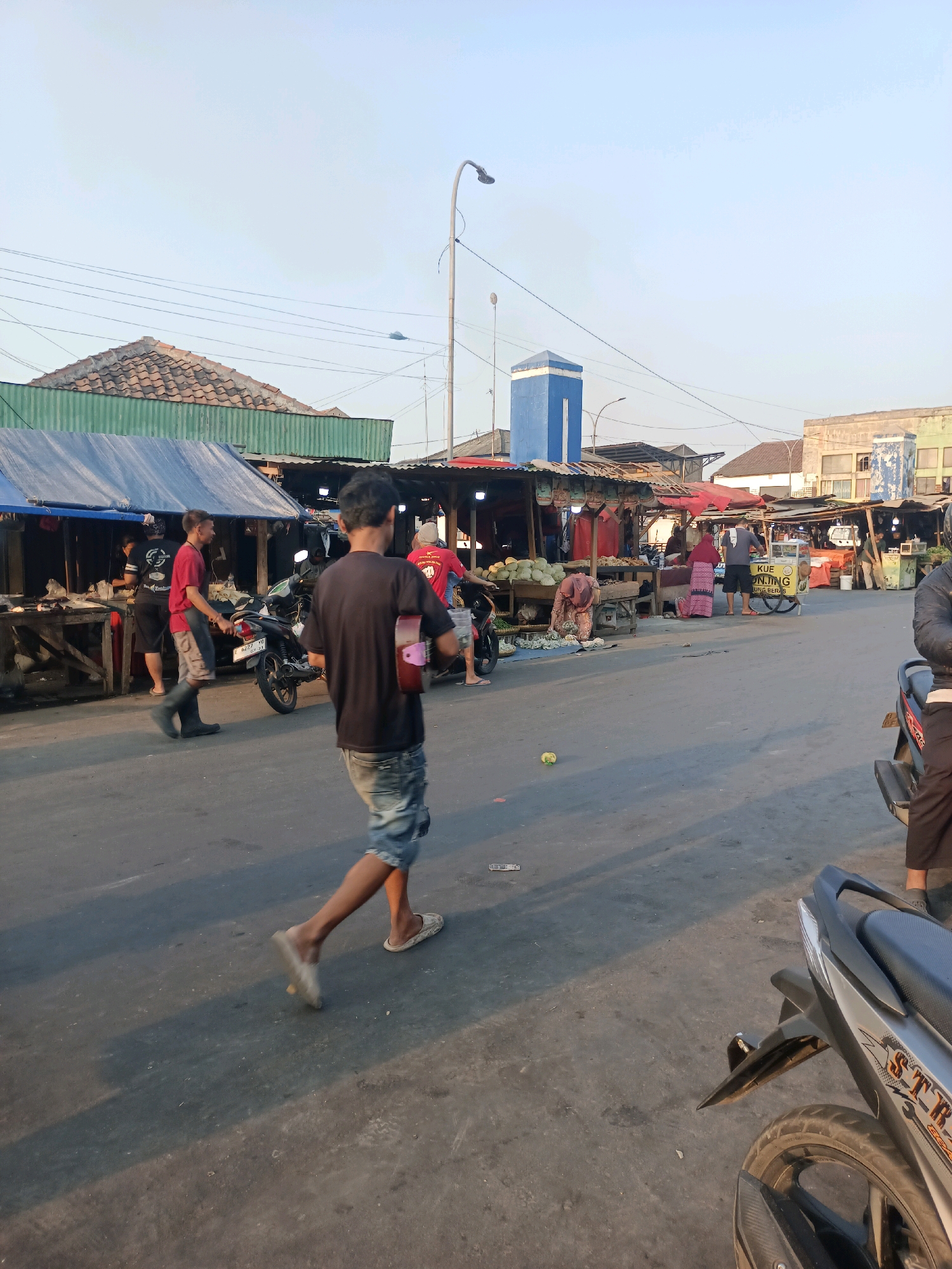 The image depicts a bustling outdoor market scene in Karawang, Indonesia. In the foreground, a young man in denim shorts and a dark t-shirt walks away from the camera, carrying a pink object that resembles a small case. To his left, another man in a red shirt and black boots walks towards a parked motorcycle. Behind them, several market stalls are set up with awnings and goods displayed. One stall prominently features a yellow sign with Indonesian writing, possibly advertising food or goods. People are seen moving between stalls, interacting with vendors, and operating motorcycles. The environment appears to be a street or open area with concrete pavement. Buildings, including a distinctive blue tower, are visible in the background, along with overhead power lines and a street lamp. The lighting suggests it is either early morning or late afternoon, with long shadows cast on the ground. The market is actively operating, with a mix of individuals engaged in shopping, selling, and general movement.