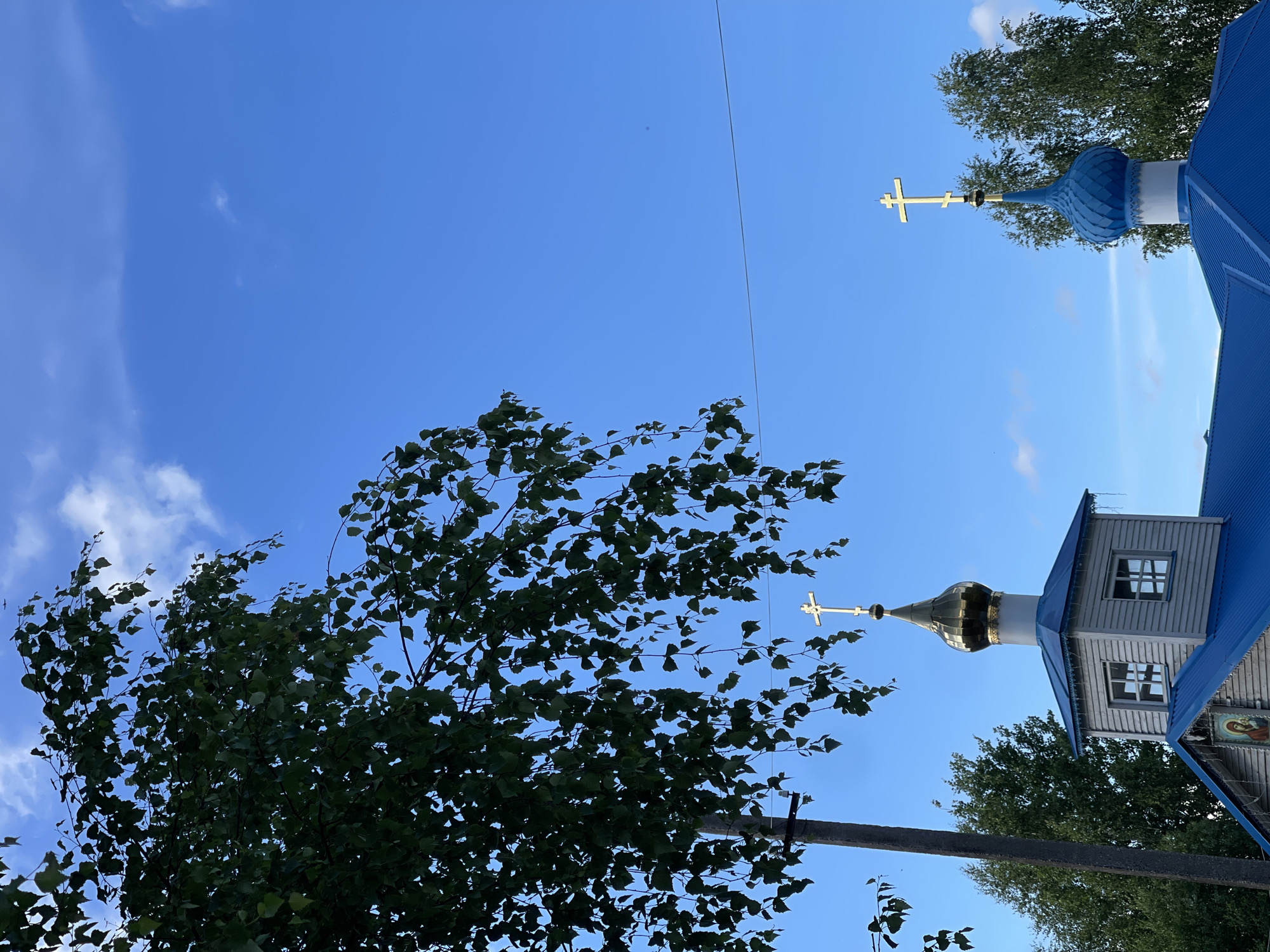 The image shows a bright blue sky with scattered white clouds. In the foreground, the branches of a leafy green tree hang from the upper left. Two church domes, one golden and one blue, are visible against the sky, each topped with a gold cross. The golden dome is part of a grey wooden structure with white-trimmed windows. The blue dome is part of a building with a blue metal roof. The building is partially obscured by the tree. The overall scene appears to be a peaceful, sunny day in a rural or suburban setting, likely in or near the city of Zarechny, Russia, given the architectural style of the church. There are no people visible in the image.