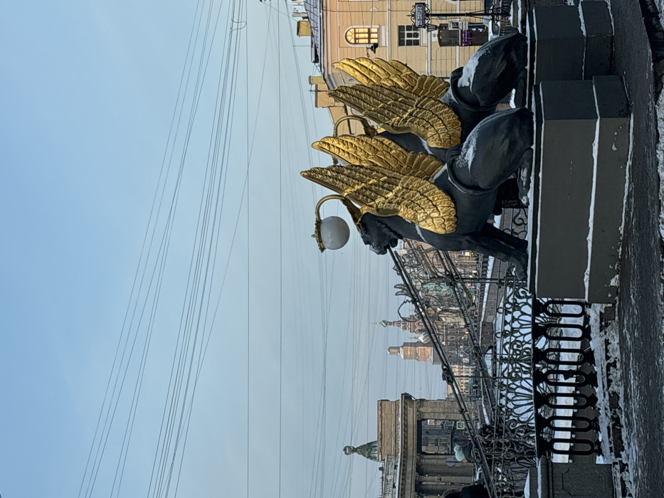 The image captures a winter scene on the Bank Bridge (Bankovsky Bridge) over the Griboyedov Canal in Saint Petersburg, Russia. In the immediate foreground, two dark-colored winged griffin statues with distinct golden wings sit on rectangular pedestals, with snow lightly dusting their backs and the ground around them. A white spherical lamp on a curved fixture extends from the back of the griffin closest to the viewer.
Behind the statues, the intricate black metal railings of the bridge are visible, also partially covered with snow. Several pedestrians, mostly indistinct, are seen walking across the bridge in the mid-ground. To the far left, a classical building featuring columns and a small, greenish domed structure is partially visible. Centered in the background, the ornate, multi-domed architecture of the Church of the Savior on Spilled Blood is clearly identifiable. To the right, a light-colored building with multiple windows stands adjacent to the bridge. The sky is a pale blue, and numerous thin electrical wires crisscross the upper portion of the frame. The scene is static, documenting a cold, clear urban day.