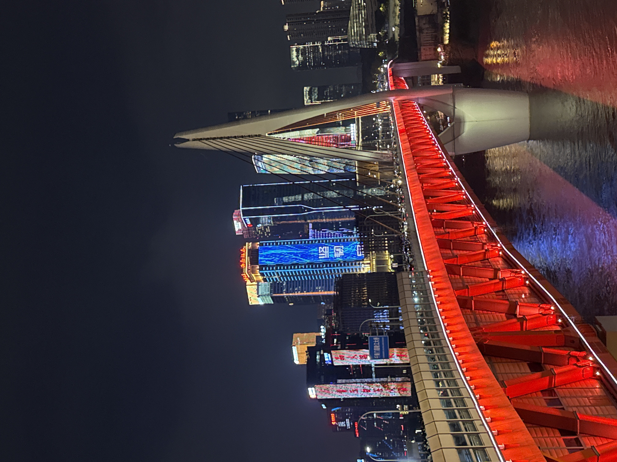 A nighttime, high-angle, long shot captures a brightly lit cable-stayed bridge spanning a river in Yuzhong District, China. The bridge, a prominent structure in the foreground, is illuminated with vibrant red lights on its underside and along its railing, casting a warm glow onto the dark water below. The bridge's suspension cables and a portion of its tower are visible. In the background, a cluster of modern skyscrapers rises, adorned with a variety of colorful and dynamic light displays, including blue and white patterns and vertical strips of light. A road with visible traffic runs along the bridge, and streetlights are also present. The overall scene depicts a bustling urban environment at night, characterized by extensive artificial illumination from both the bridge and the surrounding city architecture.