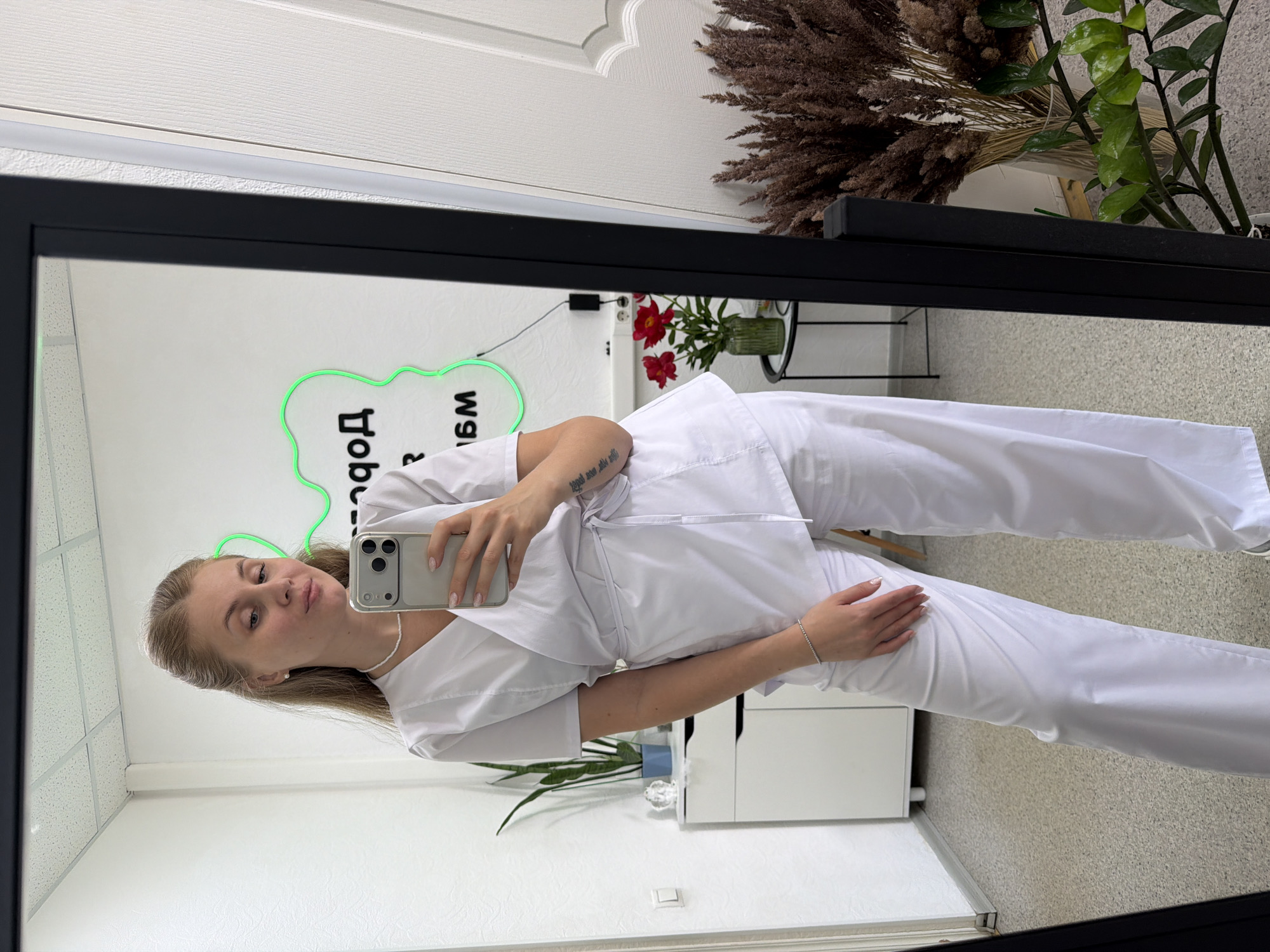 A young woman is captured in a mirror selfie, standing sideways and holding a silver smartphone with her right hand to take the picture. She has light brown hair, styled back from her face, and wears a pristine white medical scrub top and matching pants. Her left hand rests on her hip. She accessorizes with a white beaded necklace, a small earring, a silver bracelet on her right wrist, and light-colored nail polish. A tattoo is visible on her left forearm. Her expression is calm and composed.
The reflection in the large, black-framed mirror reveals a clean, professionally organized interior. The walls are white, with one section appearing textured. The ceiling is fitted with white acoustic tiles. Decorative elements include a large bouquet of dried pampas grass or similar reeds, a vibrant green leafy plant, and a vase containing red flowers on a surface behind her. A prominent green neon sign on the wall displays text which, when un-mirrored, reads "Добро" (Dobro), likely part of "Добро пожаловать" (Welcome) in Russian. A white cabinet or desk with drawers is also visible in the background. The scene suggests a professional environment, such as a medical office, clinic, or aesthetic salon in Chaikovsky, Russia.