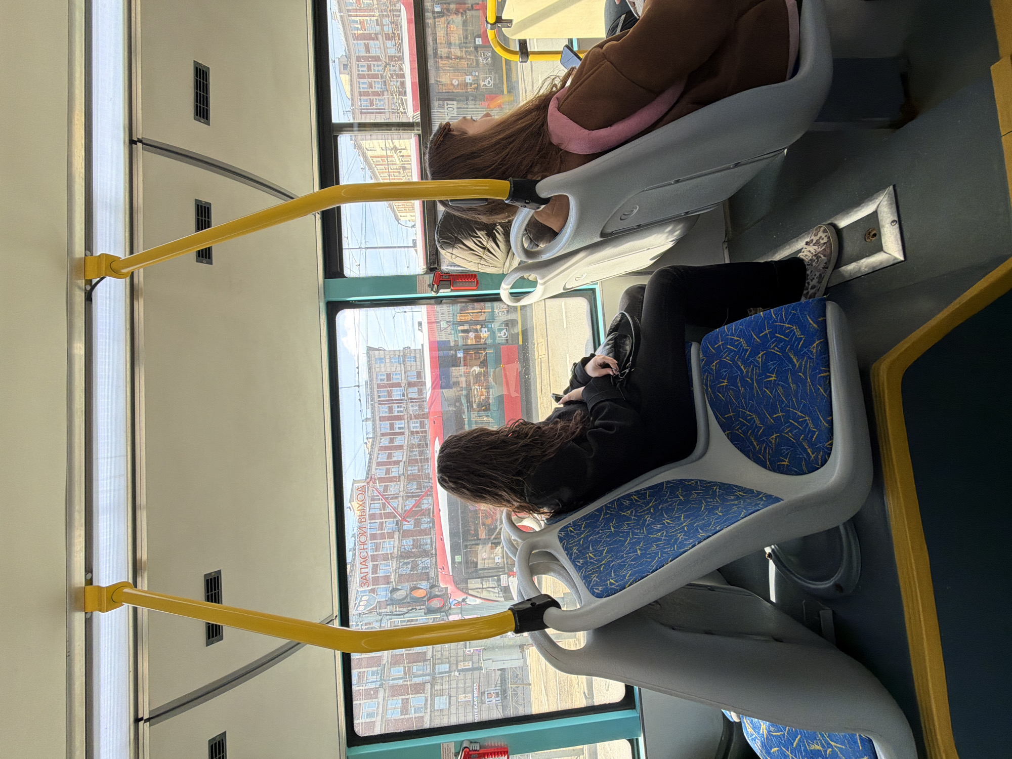 Two women are seated on a bus. One woman, wearing a brown coat and a pink neck pillow, is seated in the forward-facing seat. The other woman, wearing a black top and black pants, is seated in the rear-facing seat. Both seats are blue and gray. The bus has yellow handrails. Through the window, buildings are visible, including text in Russian that identifies the location as Saint Petersburg, Russia.