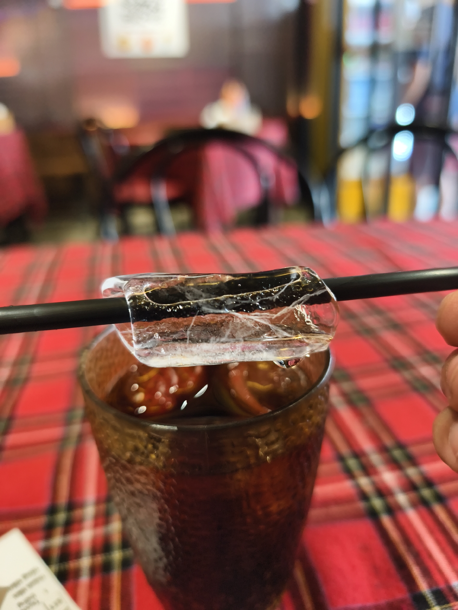 A close-up shot shows a dark-colored glass filled with liquid and ice, a black straw inserted through the ice, and the edge of a hand visible in the right side of the frame. The table below is covered in a red plaid tablecloth. In the background, blurry objects and people suggest a restaurant setting. The image was taken in Bac Nha Trang Ward, Vietnam.