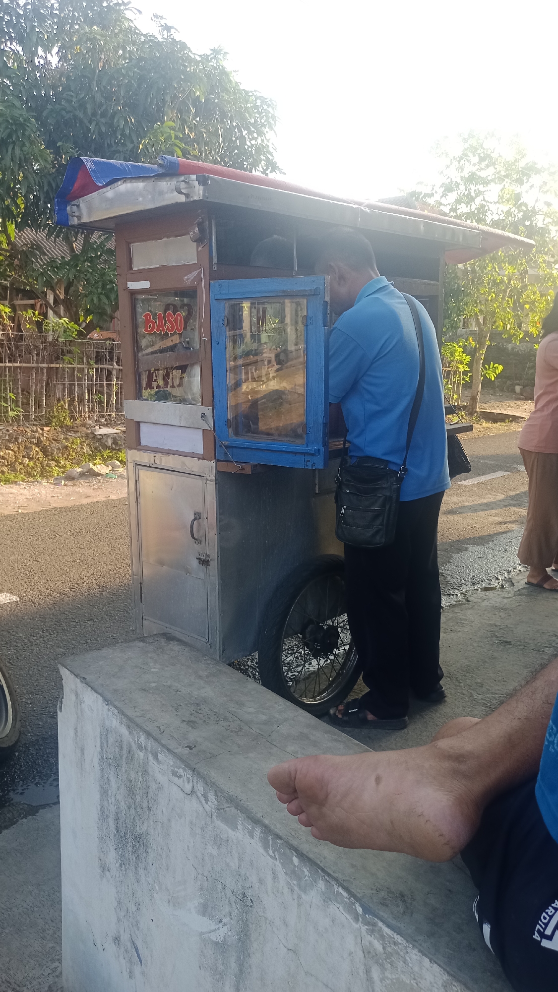 In Pakemitan, Indonesia, a man wearing a blue shirt and black pants stands at a mobile food cart labeled "BASO." He is looking into the cart, which has a blue-framed glass window. A black shoulder bag is slung across his chest. To the right, a person in a pink shirt and brown pants stands near the cart. In the foreground, a bare foot rests on a concrete structure. The food cart is on a paved surface, with vegetation and trees in the background. The sun is shining, casting a bright light.