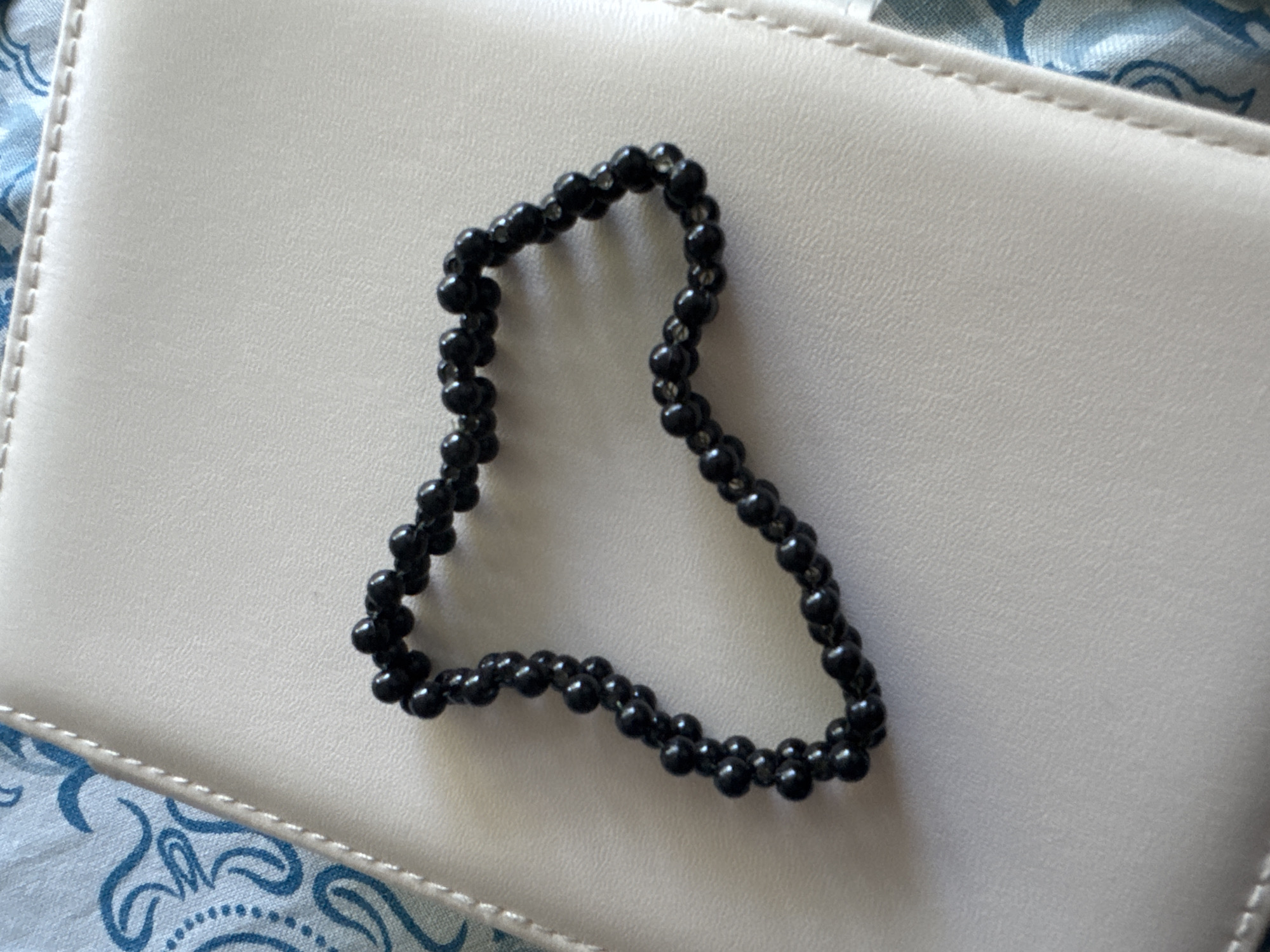 A black beaded bracelet is laid out on a closed, light-colored book or notebook. The bracelet is made of small, round black beads, and it appears to be elasticated, as it is not a perfect circle but has a more organic, slightly irregular shape. The book has a textured surface and a visible seam along its edge. The background to the left of the book shows a blue and white patterned fabric, possibly a tablecloth or scarf. The lighting is bright, casting a subtle shadow from the bracelet. The image is a close-up shot, focusing on the bracelet and the book. There is no information available about its location in the city of Яромінскі сельскі Савет, Belarus.