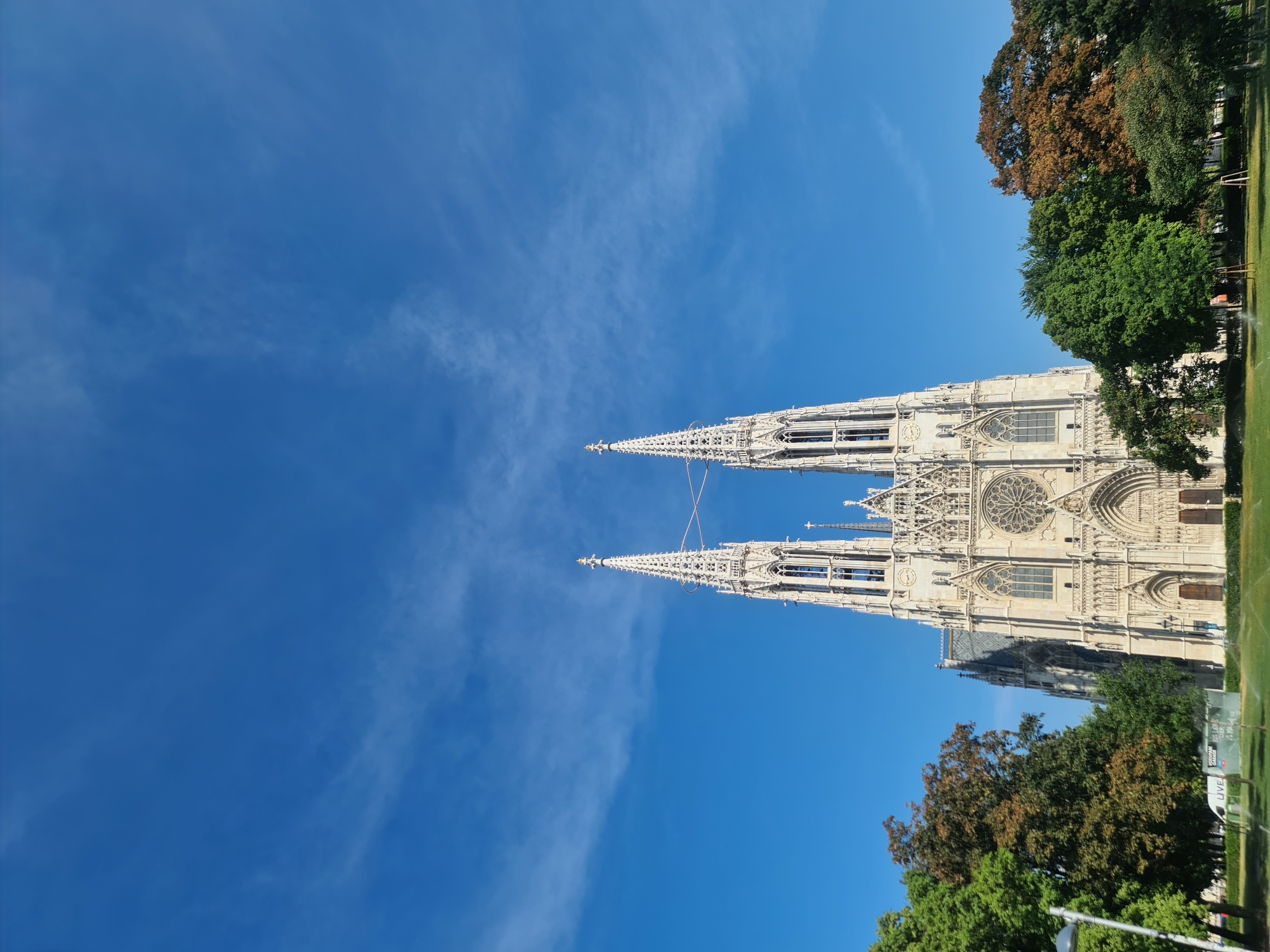 The image shows the Votivkirche, a Gothic Revival church in Vienna, Austria, seen from a slightly low angle, with its two prominent spires reaching towards a bright blue sky dotted with wispy clouds. The church's intricate stone facade, with its many windows, arches, and ornamental details, is visible in the center of the frame. In front of the church, a green lawn stretches out, with scattered trees that are mostly green, though some exhibit autumn colors. A lamppost is visible on the left side of the lawn. The overall scene depicts a clear, sunny day with the architectural grandeur of the Votivkirche as the central focus, surrounded by a park-like setting.
