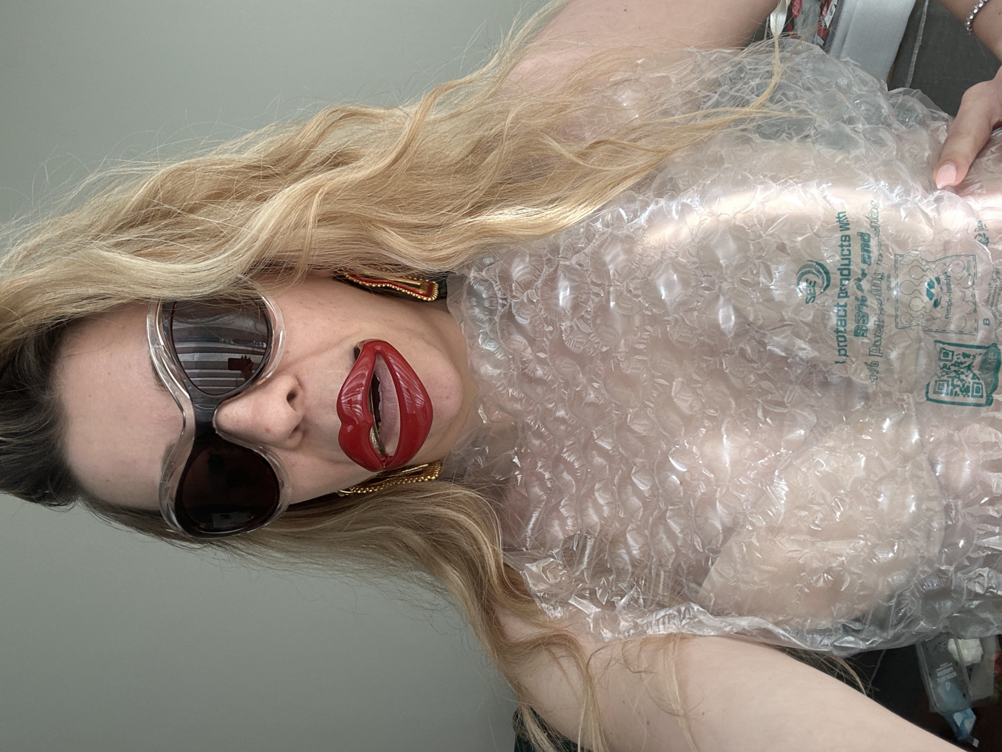 The image displays a close-up portrait of a person, likely female, with long, wavy blonde hair, presented in a rotated orientation. The subject is wearing oversized clear-framed sunglasses with dark lenses and striking red, lip-shaped earrings with gold detailing. Her visible lips are adorned with vibrant red lipstick.
The person is partially dressed in translucent bubble wrap, which appears to be used as an unconventional garment, covering her upper torso. A hand with neatly manicured light pink nails is visible on the right side of the frame, touching the bubble wrap. The background consists of a plain, light greenish-grey wall.
The activity taking place is a direct pose for the camera, possibly a selfie, emphasizing a bold and artistic or fashion-forward aesthetic through the choice of accessories and attire.
Notable details include the texture and reflective quality of the bubble wrap, as well as the unique design of the earrings. The sunglasses reflect horizontal lines.
Several pieces of text are visible printed on the bubble wrap: "I protect products with", "35% post-consumer recycled content", and "1% packaging innovation". A "Recycle-ready" symbol, a "SEE" logo, and two QR codes are also discernible on the material.