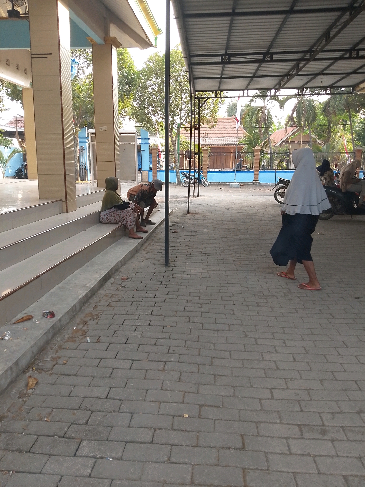 In Wadang, Indonesia, at what appears to be a community building or place of worship with a covered outdoor area, three individuals are present. Two people are seated on the steps of the building, facing away from the camera. The individual on the left, wearing a green headscarf and patterned pants, sits with their legs extended. Next to them, an individual in a brown shirt and shorts, wearing a cap, is crouched. Further into the scene, a third individual, dressed in a white headscarf and dark skirt, walks away from the camera across a paved courtyard. Beyond the courtyard, a fence encloses buildings and trees. A motorcycle is visible parked on the left side of the frame. The ground is paved with interlocking gray bricks, with some debris scattered on the steps and the paved area. The lighting suggests it is daytime.