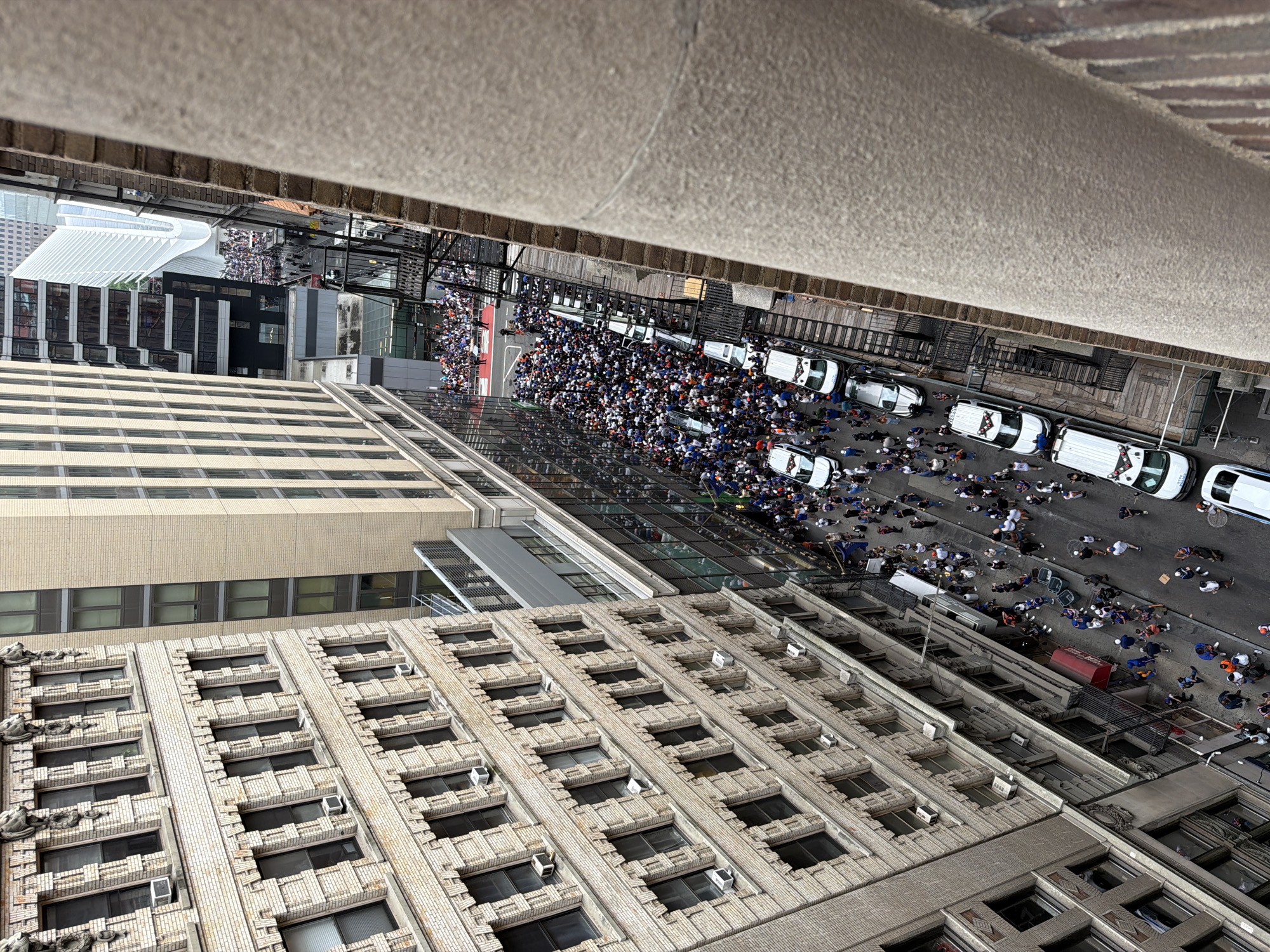 This image captures an aerial view of a street scene in a dense urban environment, likely New York City, given the architectural style and layout. The primary subjects are a large crowd of people gathered on the street and sidewalks, interspersed with several white police vehicles. The people are dressed in casual clothing, with many wearing blue shirts, suggesting a unified group or event.
The activity taking place appears to be a parade or a large public gathering, possibly a celebration or demonstration, given the presence of police escort. The crowd is dense, filling much of the available street space, and they are looking in various directions, some towards the buildings, others towards the street ahead. The police vehicles are positioned along the street, seemingly managing the crowd and traffic flow.
Notable details include the time of day, which appears to be daytime with overcast skies or the direct overhead sun casting minimal shadows. The buildings in the foreground and midground are tall and modern, with glass facades and industrial-style brickwork, typical of a major city. In the background, more modern structures, including what appears to be the Oculus transportation hub, are visible, further solidifying the New York City context. The mood seems to be one of excitement or anticipation, with the large gathering suggesting a significant event. There is no visible text in the image.