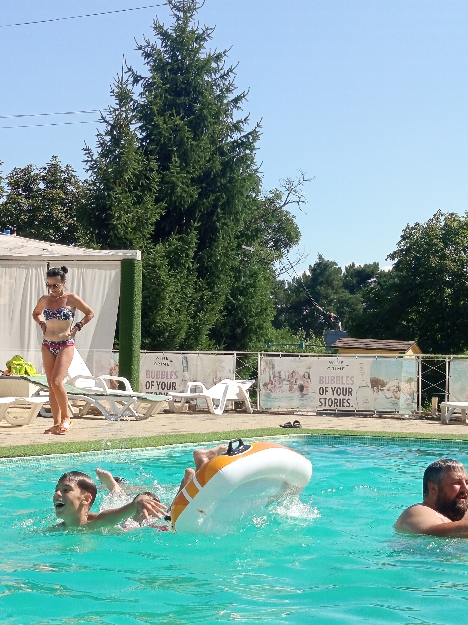 The image shows a sunny day at an outdoor swimming pool facility. In the foreground, two boys are in the bright turquoise water. The boy on the left is laughing and splashing, while another boy is partially visible behind him. An inflatable toy, white with orange accents, is partially submerged and creating splashes. To the right, a man with a beard is in the water, looking to his right. In the background, a woman in a patterned bikini stands on the pool deck, with her hands on her hips, wearing sunglasses and flip-flops. Behind her, lounge chairs and a covered seating area are visible, along with lush green trees and a clear blue sky. A banner displaying text and images is stretched across the back of the pool area. The scene appears to be a recreational area in Molovata Nouă, Moldova.
