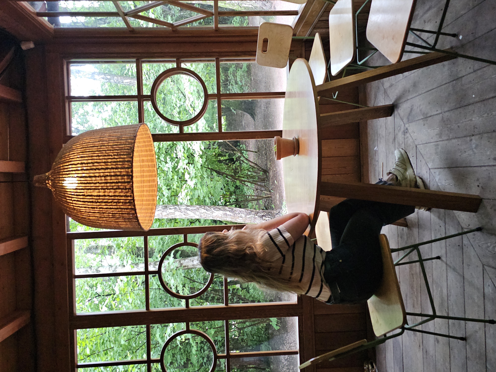 A woman with blonde hair is sitting at a round, light-brown wooden table in a room with large windows. She is wearing a striped t-shirt and dark pants. The room is decorated with wooden paneling and has a large, woven pendant lamp hanging from the ceiling. Outside the windows, there are trees and greenery, suggesting a natural or park-like setting. The floor is made of wooden planks. The location is identified as the city of Nemchinovo, Russia.