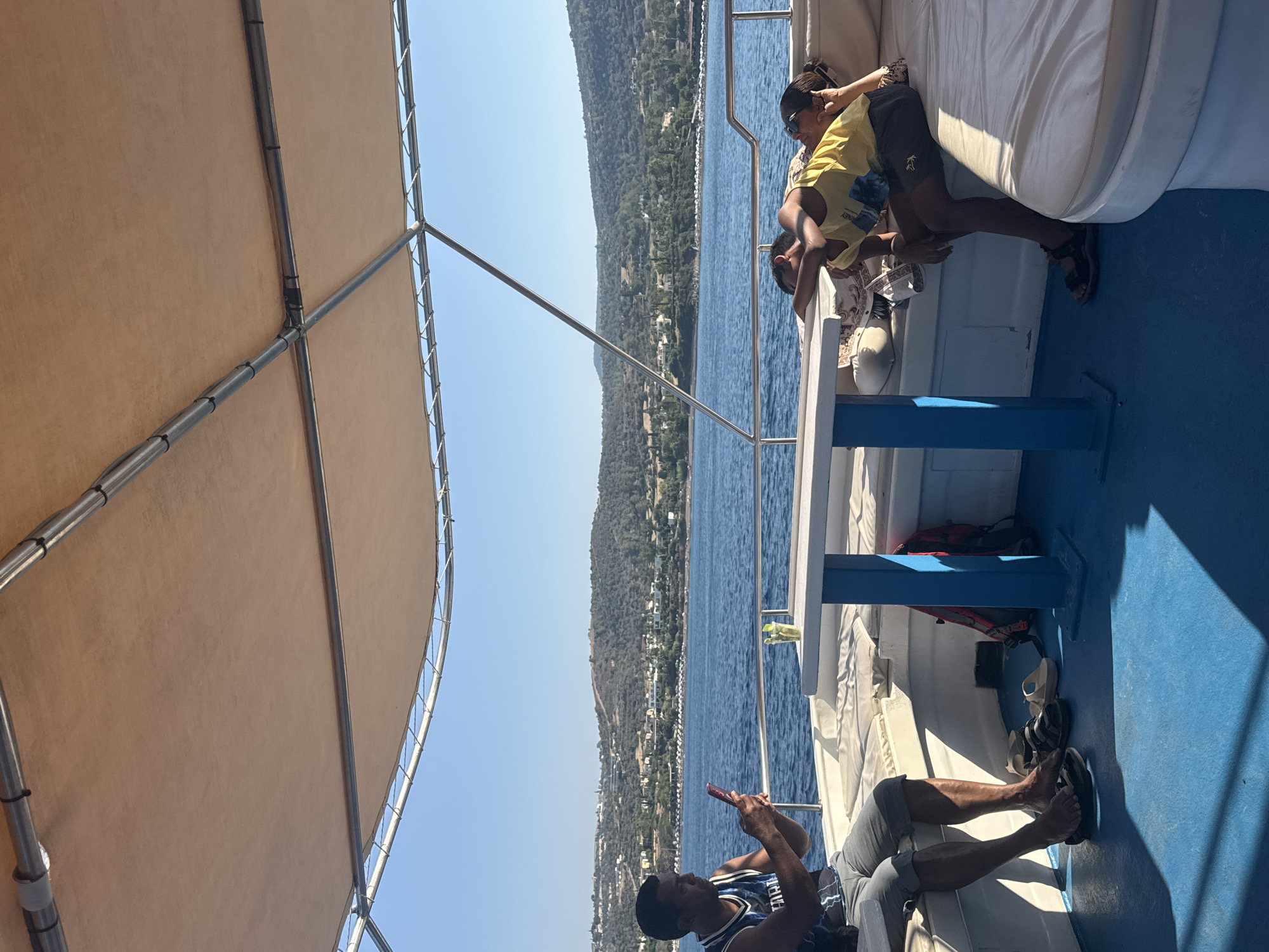 The image depicts a scene on the deck of a boat under a tan canopy. In the foreground, a man in a black and blue tank top and gray shorts sits on a white bench, holding a red smartphone and appearing to take a picture or video. His bare feet are resting on the blue deck. To his right, a white table with a blue base stands between two benches. On the table, there is a clear glass with a green beverage and ice. Further back on the right, a young person in a yellow t-shirt and dark shorts is lying down on a white padded bench, with their head resting on another person's arm. The second person, partially visible and wearing sunglasses, is lying next to them. The background shows a calm blue sea and a hilly coastline with buildings and trees, suggesting a location near Neo Chorio, Cyprus. The lighting indicates a sunny day.