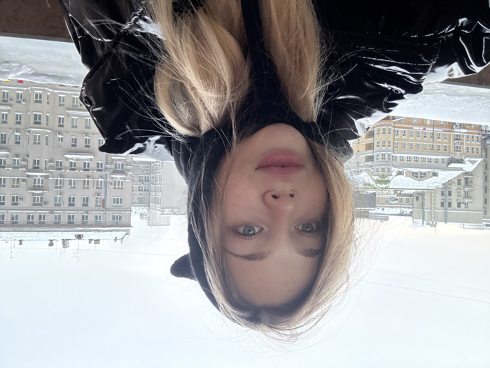A woman in a shiny black puffer jacket with a hood poses for a selfie against a backdrop of snow-covered urban buildings. The photo is taken at an unusual angle, as if the camera is held upside down. The buildings have a variety of architectural styles and are covered in snow. The sky is overcast, and the overall color palette is muted. The woman has light hair and fair skin.