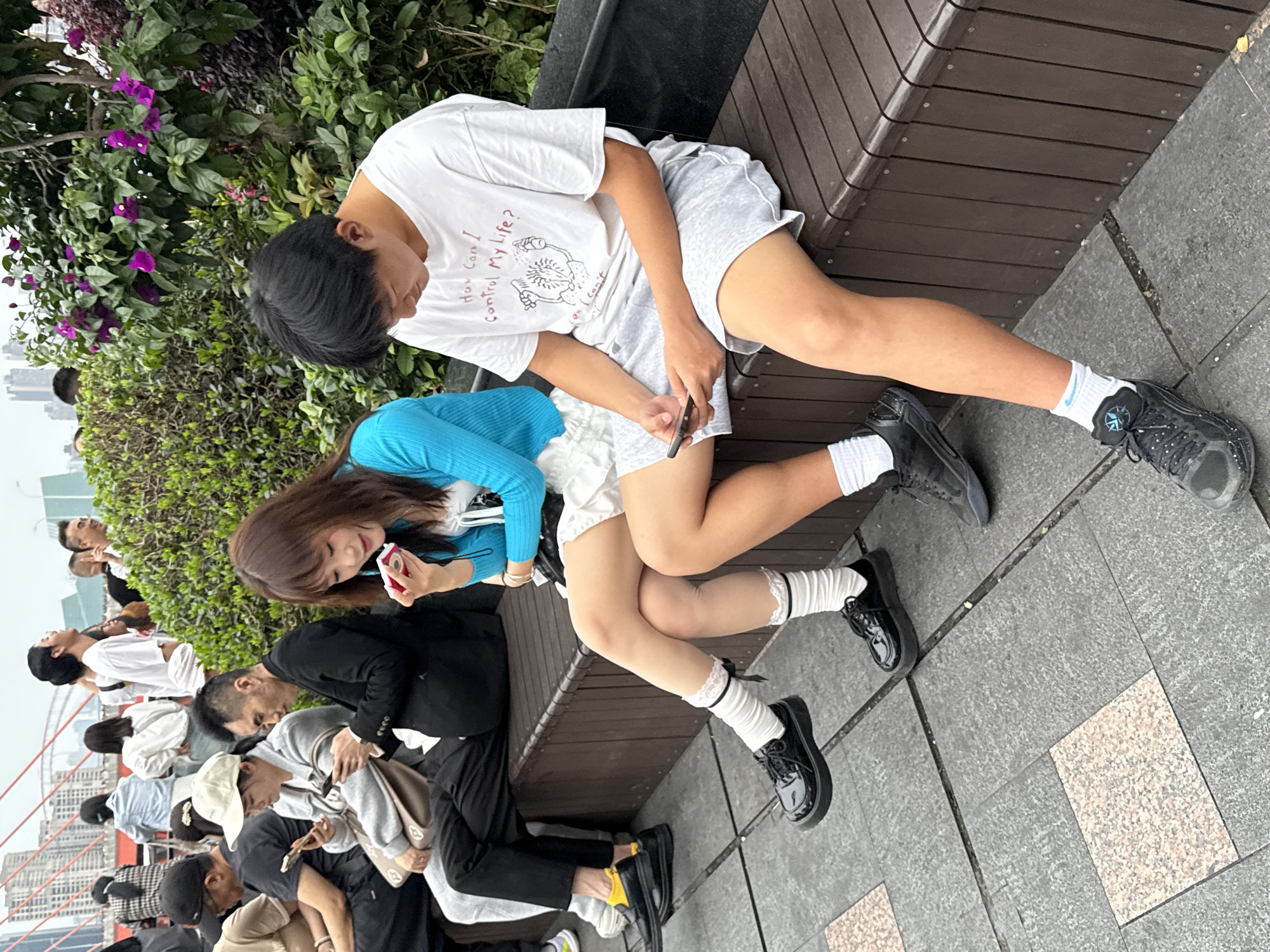 A medium shot depicts a street scene in the Yuzhong District of China. The shot is taken at eye level, and the camera is rotated counterclockwise. In the foreground, a young man and a young woman are sitting on a dark brown bench. The man is wearing a white T-shirt with a graphic and the words "How Can I Control My Life?", grey shorts, white socks, and black sneakers. He is holding a smartphone in his hands. The woman is wearing a bright blue cardigan, a white skirt, white socks with lace trim, and black patent leather shoes. She is holding a pink smartphone in her hand. They are facing each other. In the background, there are several other people seated on the bench. A variety of clothing is visible, and some individuals are holding cell phones. Green shrubbery is visible behind the bench. In the far background, buildings can be seen through the haze. The bench sits on a gray concrete surface.