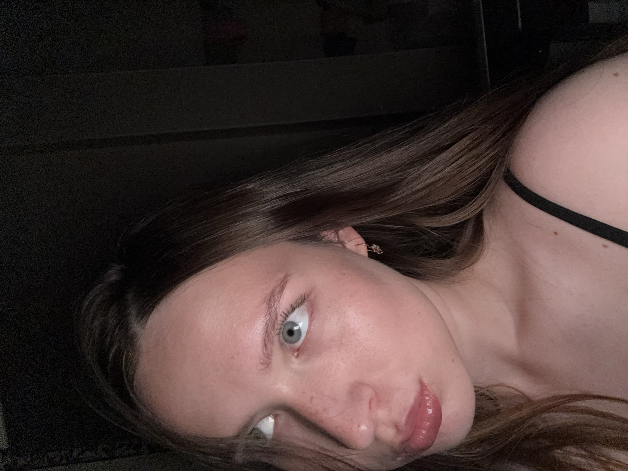 The image is a close-up portrait of a young woman with light skin, light blue eyes, and long brown hair. She is looking upwards and to her left, with her lips slightly parted. She is wearing a black strap, likely from a tank top or bra. A small star-shaped earring is visible in her left ear. The background is dark and out of focus, making it difficult to discern any specific environmental details. No geographical location or context related to Chaikovsky, Russia, can be determined from the image itself.