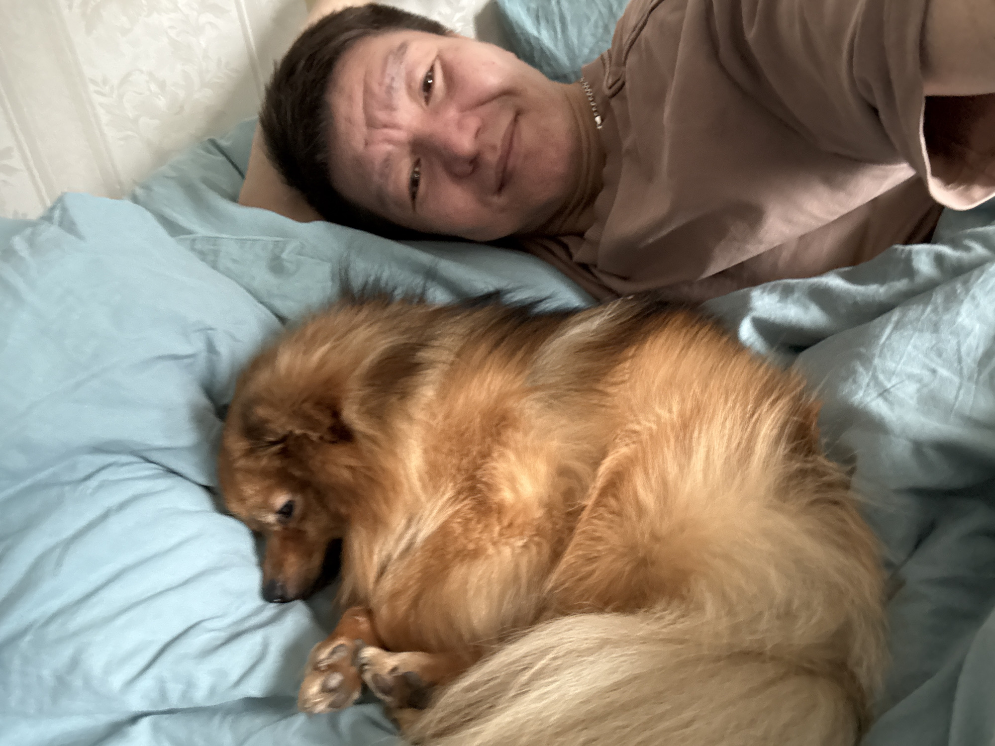 A person is lying on a bed with light blue sheets, next to a golden-brown dog curled up and sleeping. The person is fair-skinned, with short dark hair, wearing a light brown t-shirt and a silver chain. The room has light-colored patterned wallpaper visible in the background. The image appears to have been taken in a residential location, likely in Khimki, Russia, based on the user’s information. The scene depicts a moment of rest and companionship.